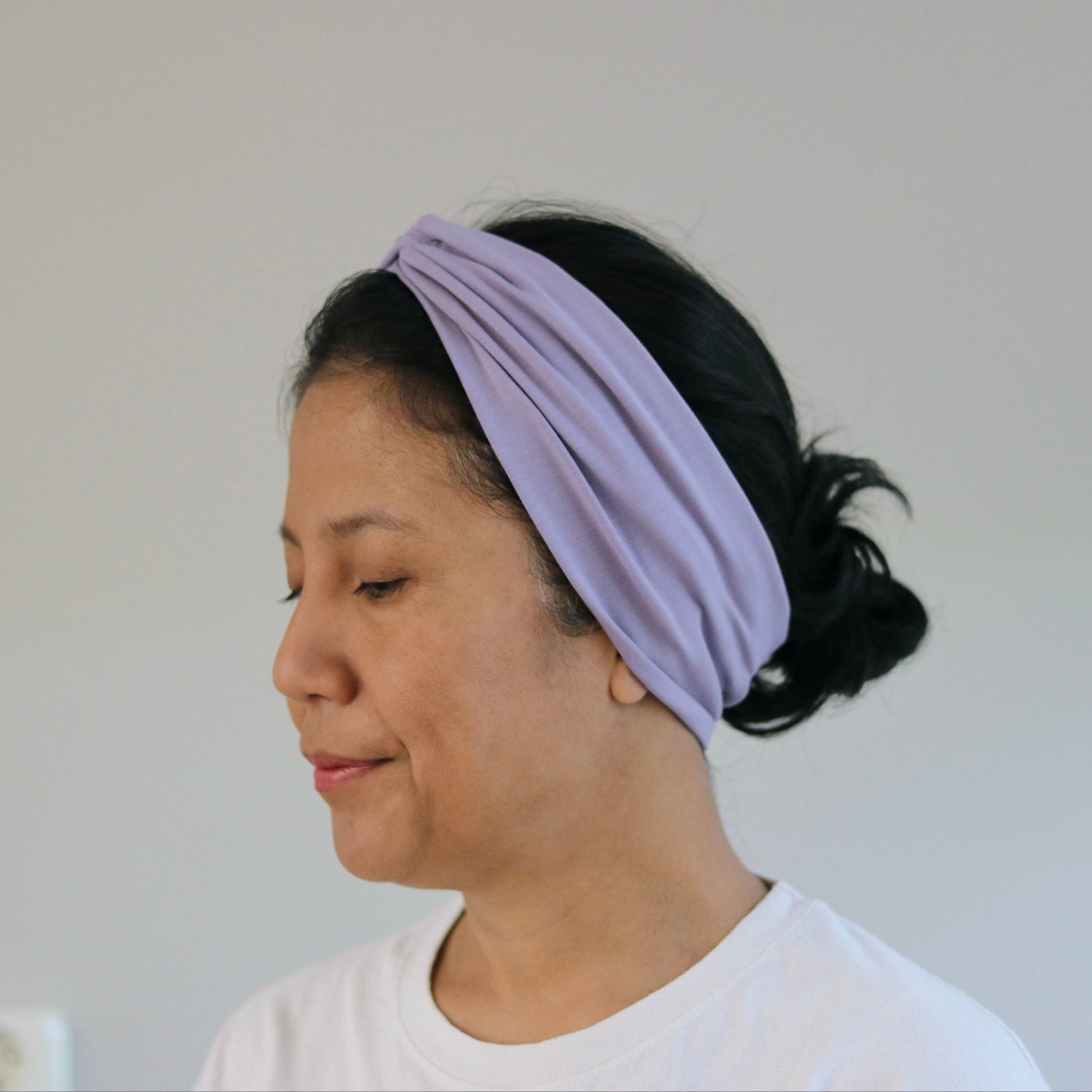 A woman wearing a light purple twisted fabric headband, shown in profile against a plain light gray wall.
