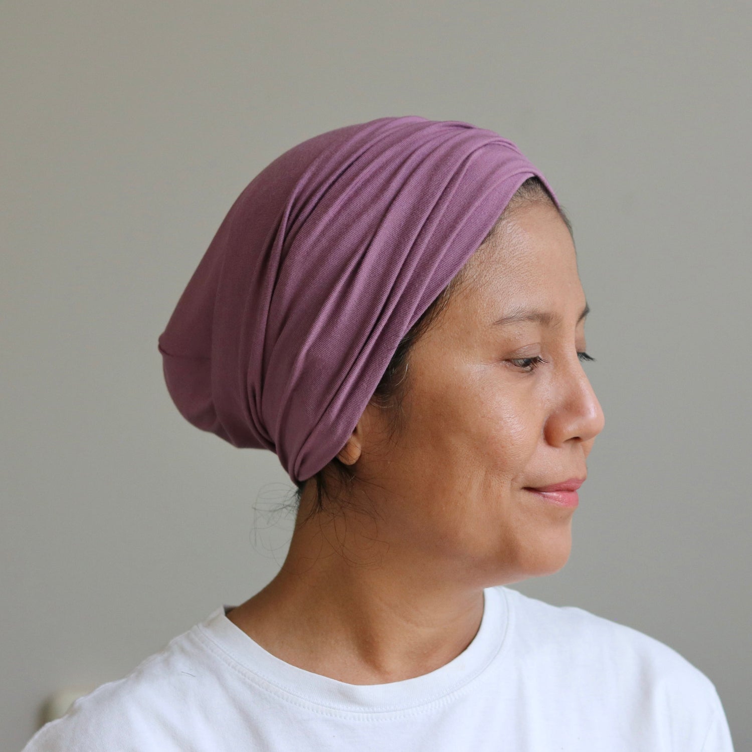 A woman in profile wearing a mauve-purple pleated fabric headwrap or hijab, paired with a white t-shirt, set against a plain light gray wall.
