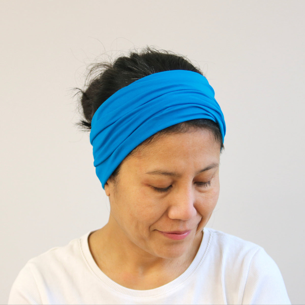 A woman wearing a bright blue knotted fabric headband and a white t-shirt, looking down against a plain light gray background.
