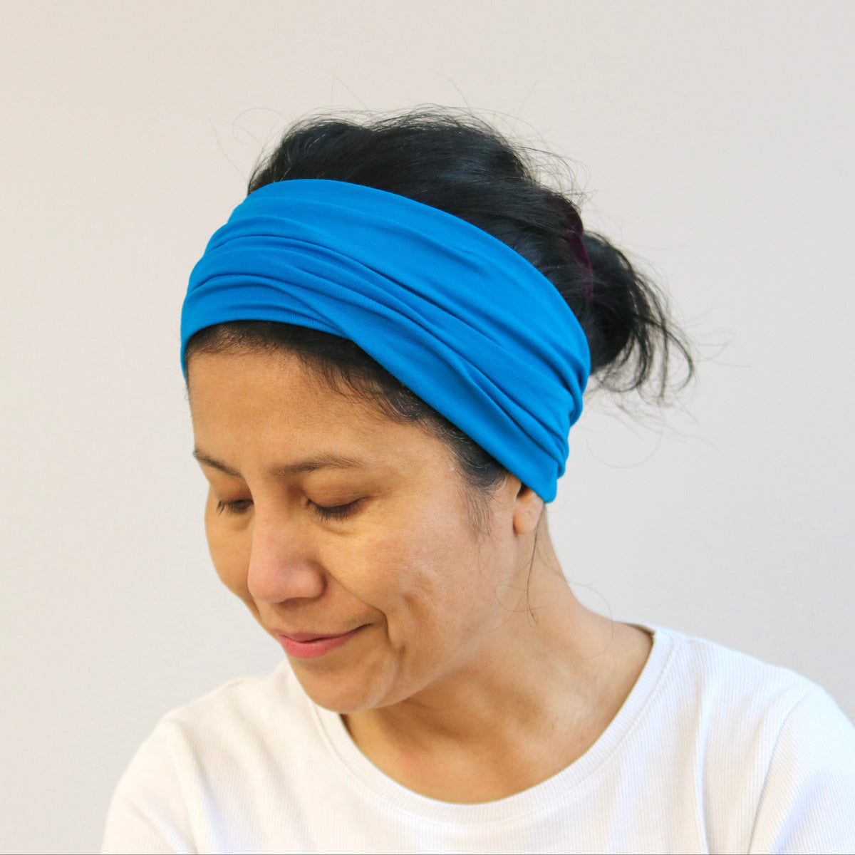A woman wearing a bright blue fabric headband and a white t-shirt, looking down against a plain light gray background.