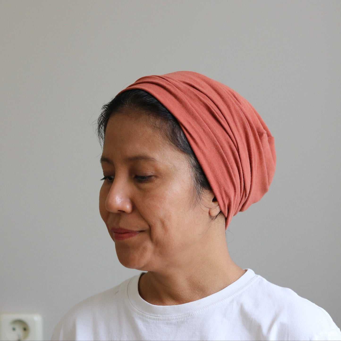 A woman wearing a terracotta-colored knitted headscarf or turban-style headwrap, paired with a white t-shirt, shown in profile against a plain light gray wall.