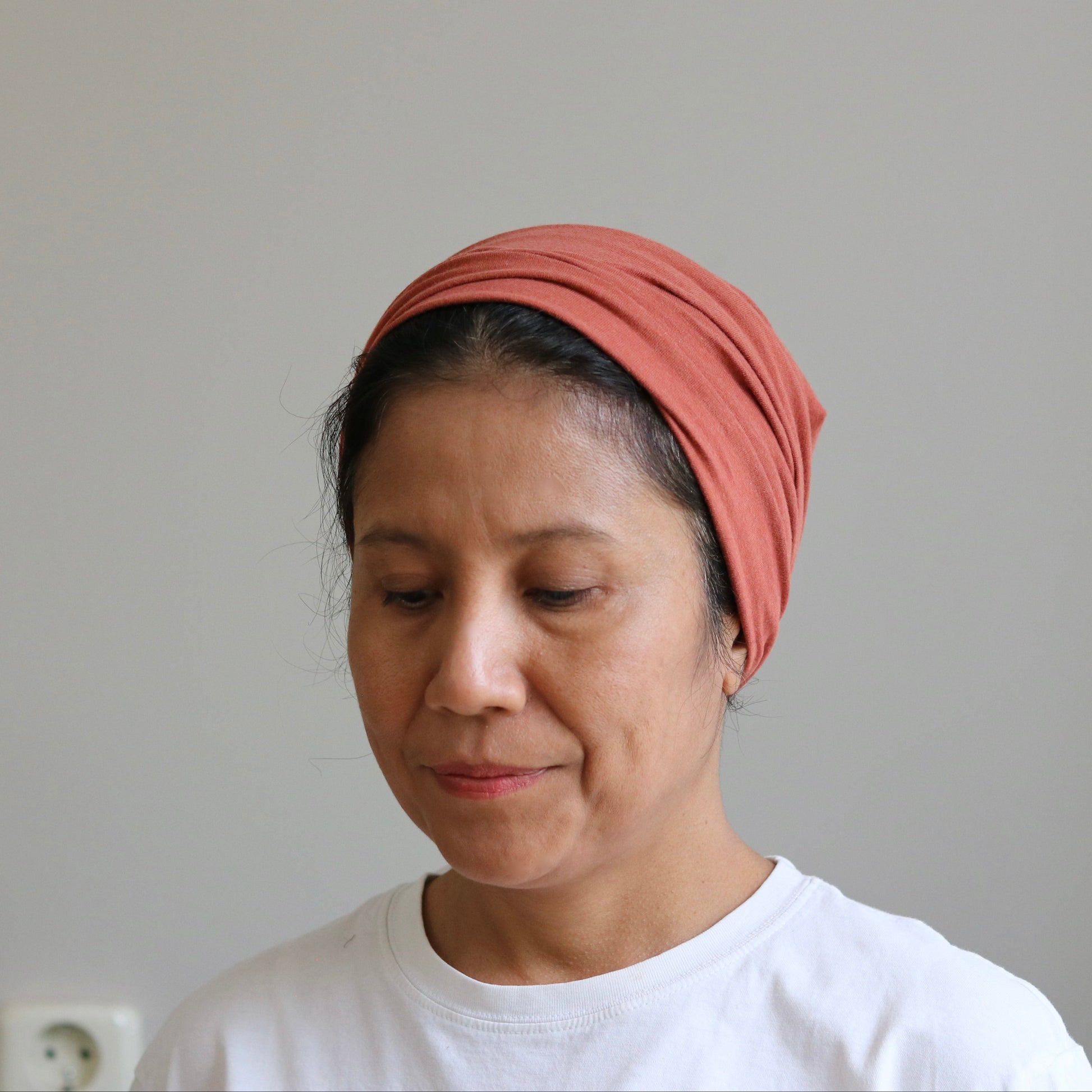 A woman wearing a dusty rose headwrap and a white t-shirt, looking down against a plain light gray wall.