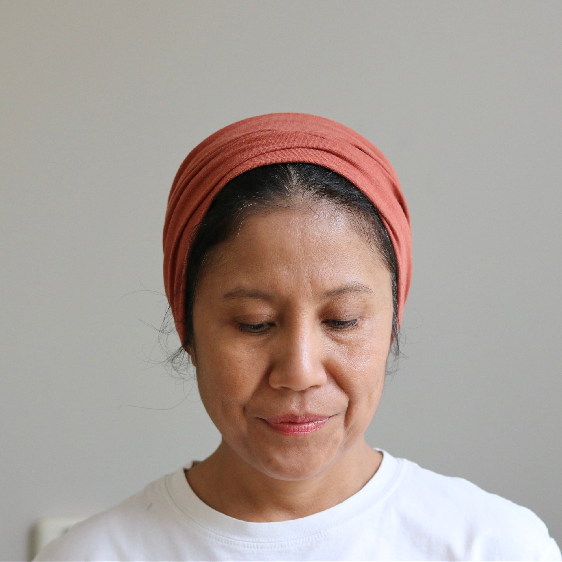 A woman wearing a terracotta-colored fabric headwrap or headband, paired with a white t-shirt, looking down against a plain light gray background.