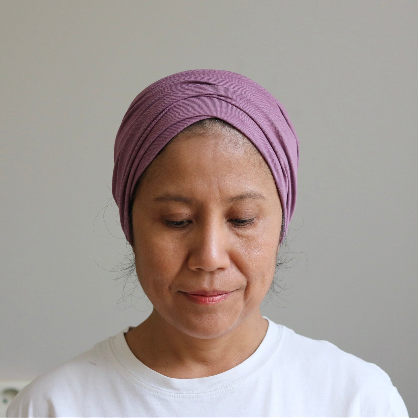 A woman wearing a mauve-purple knotted headwrap and a white t-shirt, looking down against a plain light gray wall.