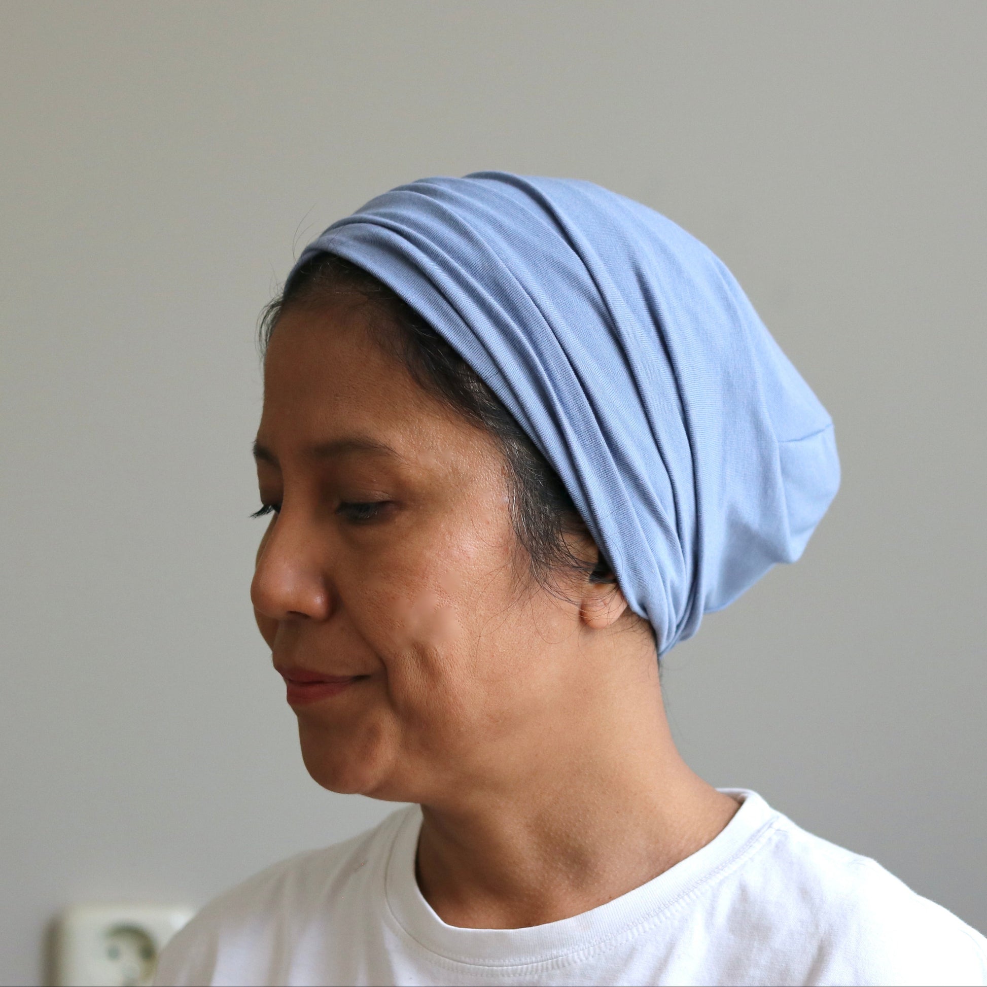 A woman wearing a light blue knotted headscarf or hijab, paired with a white t-shirt, shown in profile against a plain light gray wall.