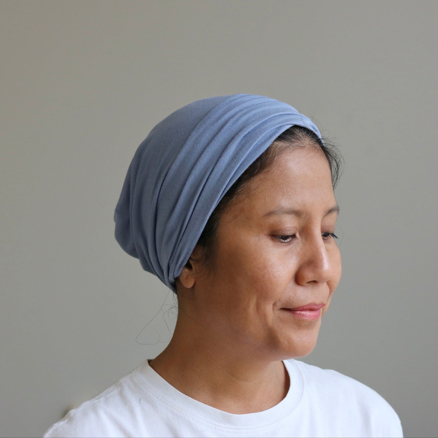 A woman wearing a light blue fabric headwrap or turban-style hair covering, paired with a white t-shirt, shown in profile against a plain light gray wall.