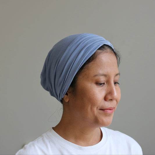 A woman wearing a light blue fabric headwrap or turban-style hair covering, paired with a white t-shirt, shown in profile against a plain light gray wall.