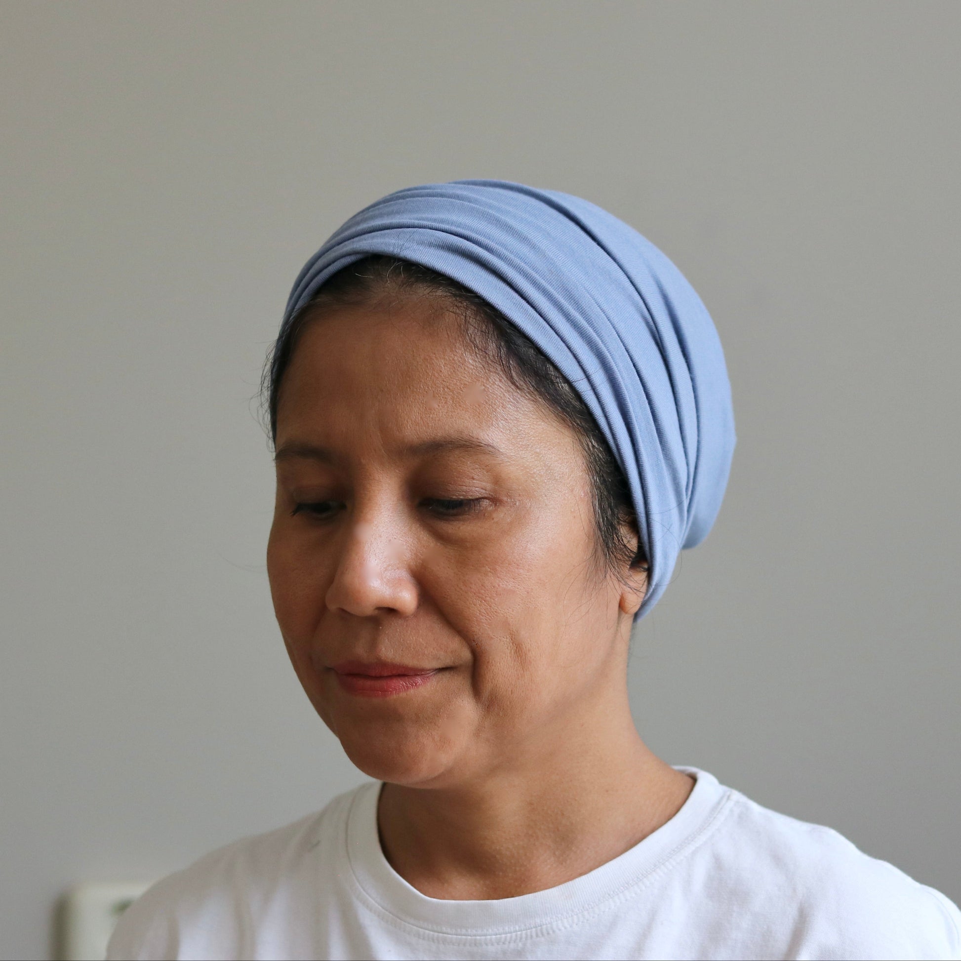 A woman wearing a light blue fabric headwrap or turban-style hair covering, paired with a white t-shirt, shown against a plain light gray wall.