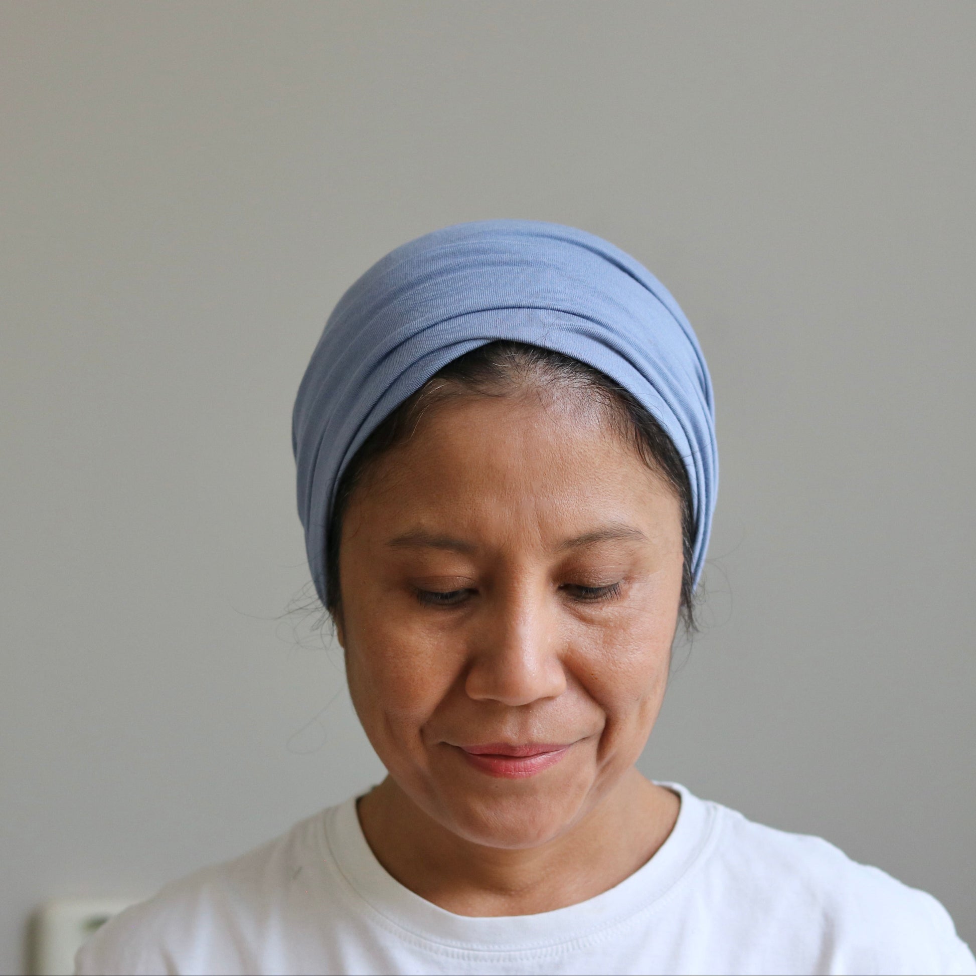 A woman wearing a light blue fabric headwrap turban and a white t-shirt, looking down against a plain light gray wall.