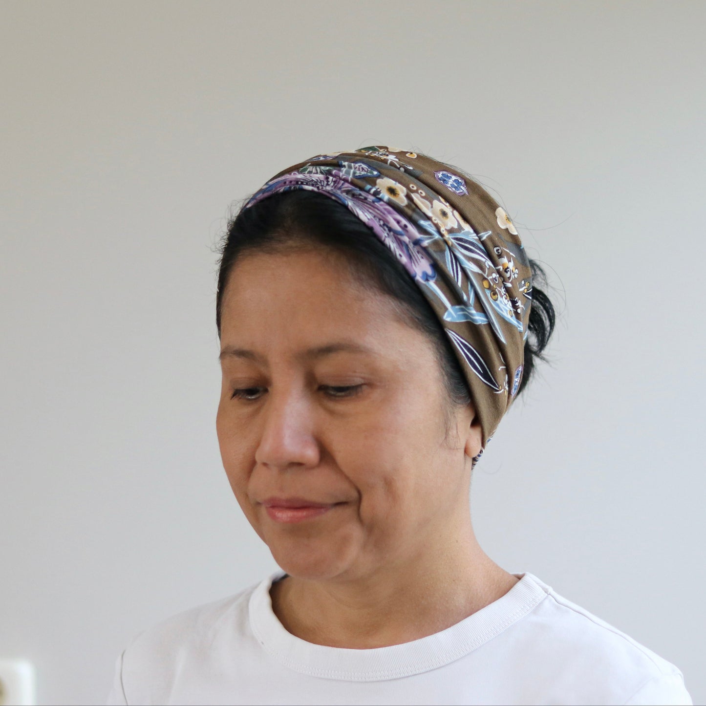 A woman wearing a patterned brown headscarf and a white t-shirt, looking down against a plain light gray wall background.