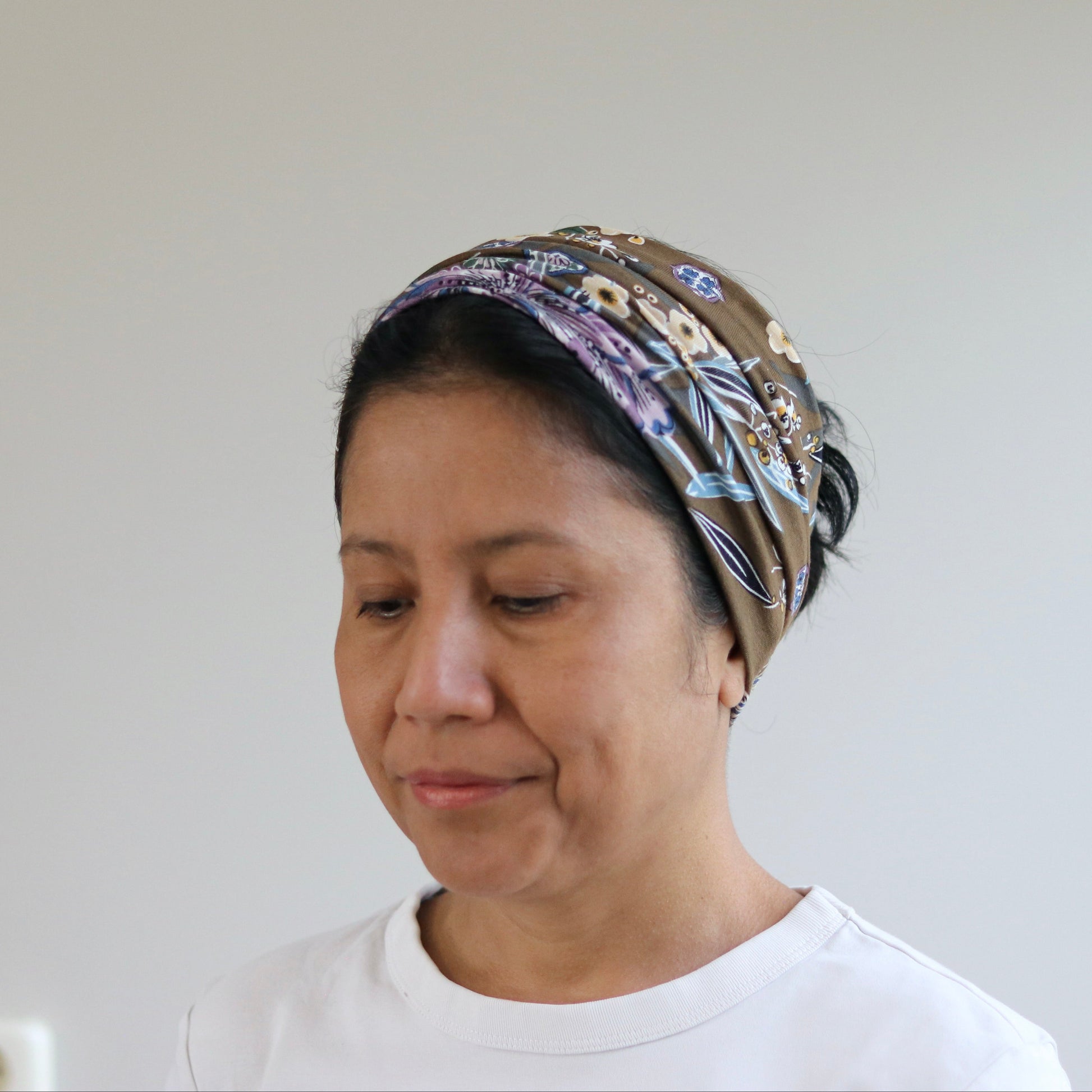 A woman wearing a patterned brown headscarf and a white t-shirt, looking down against a plain light gray wall background.