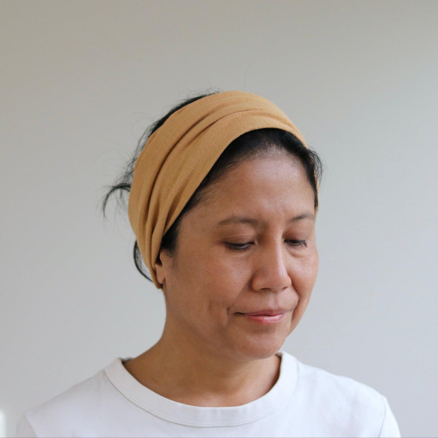 A woman wearing a caramel fabric headband and a white t-shirt, looking down with a neutral expression against a plain light gray wall.