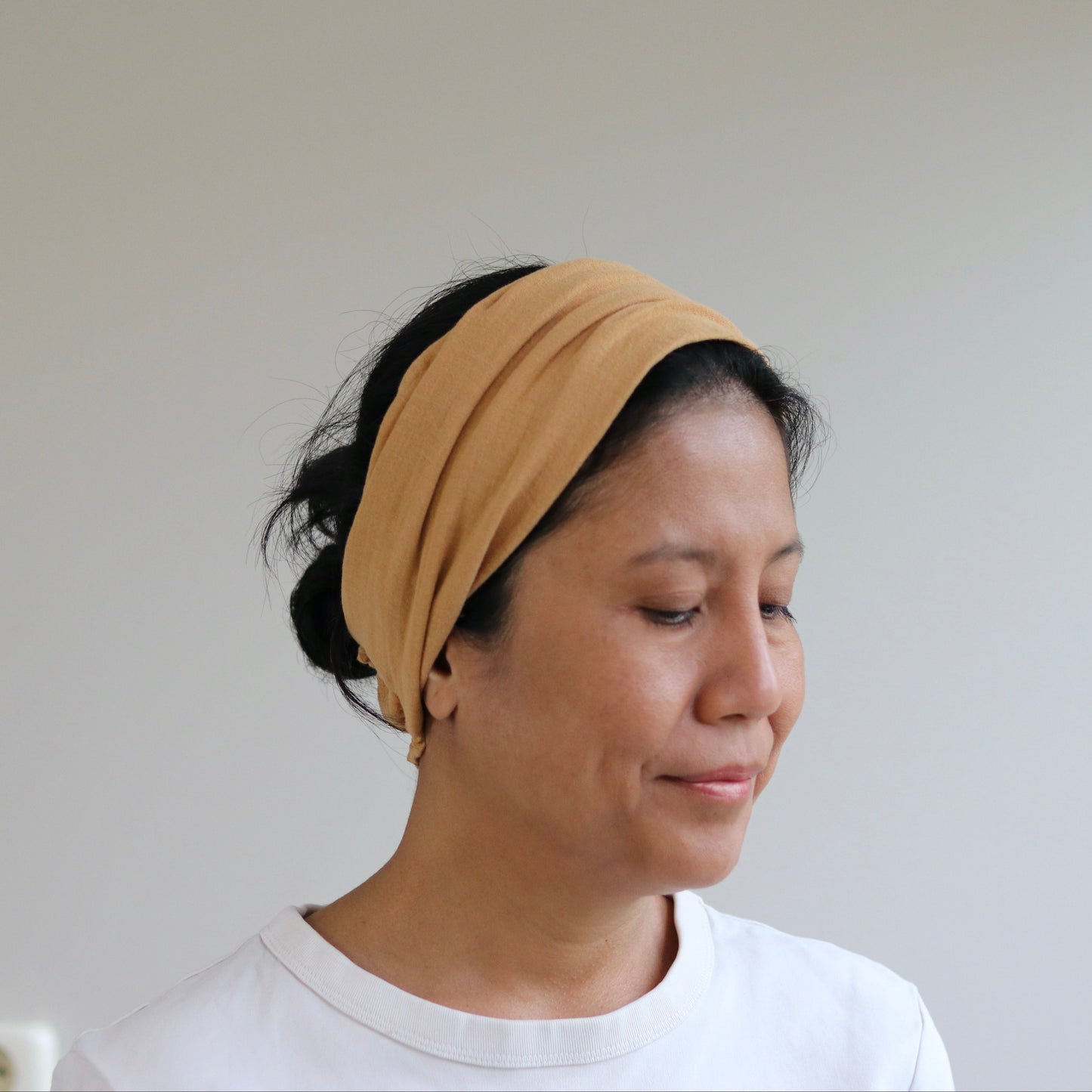 A woman wearing a caramel fabric headband and a white t-shirt, shown in profile against a plain light gray wall.