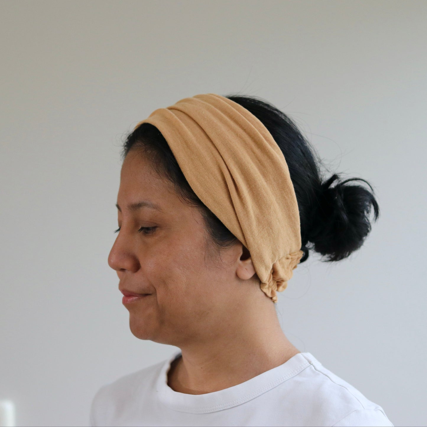 A woman in profile wearing a caramel fabric headband and a white t-shirt, standing against a plain light gray wall.