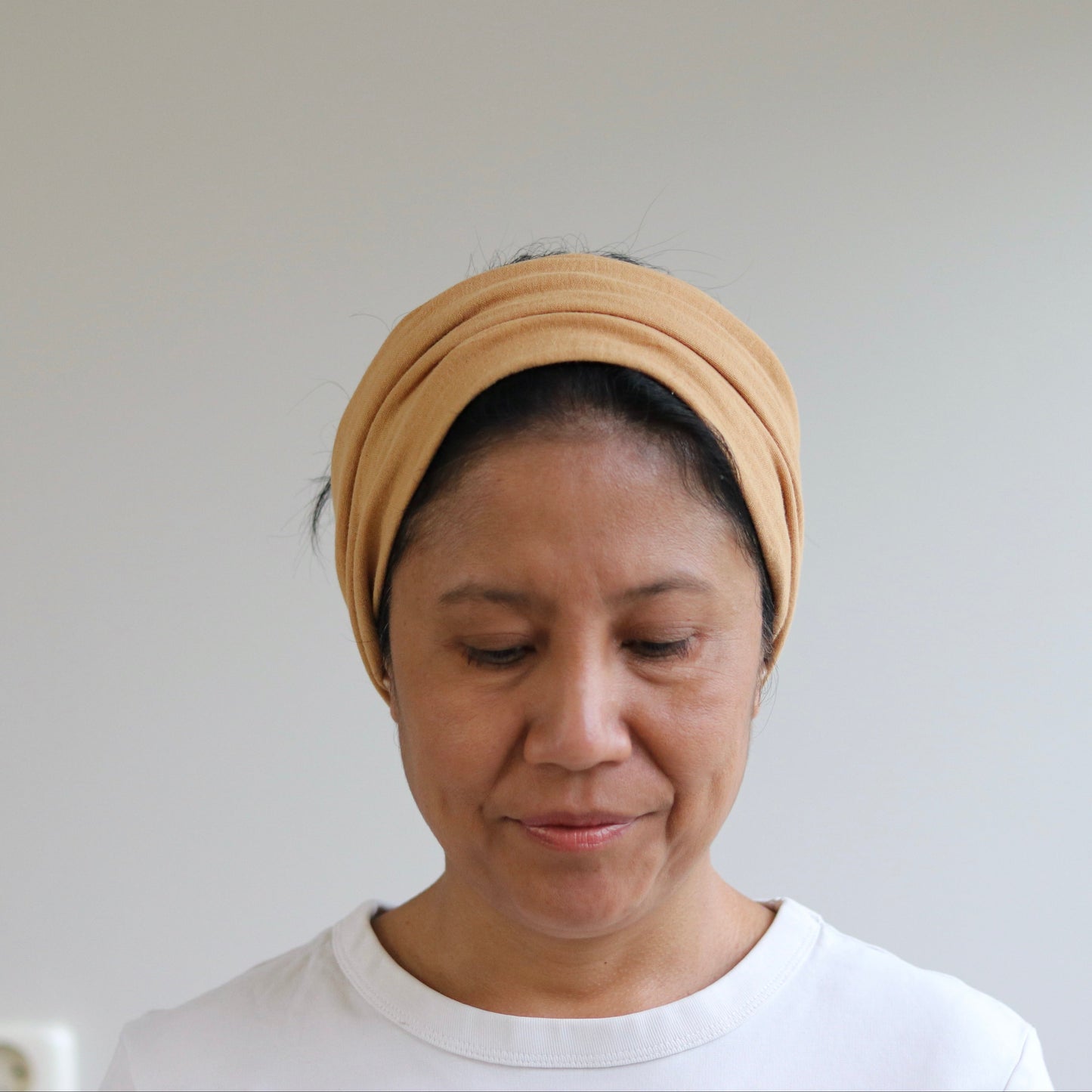 A woman wearing a caramel fabric headwrap or turban-style headband, paired with a white crewneck t-shirt, looking down against a plain light gray wall.