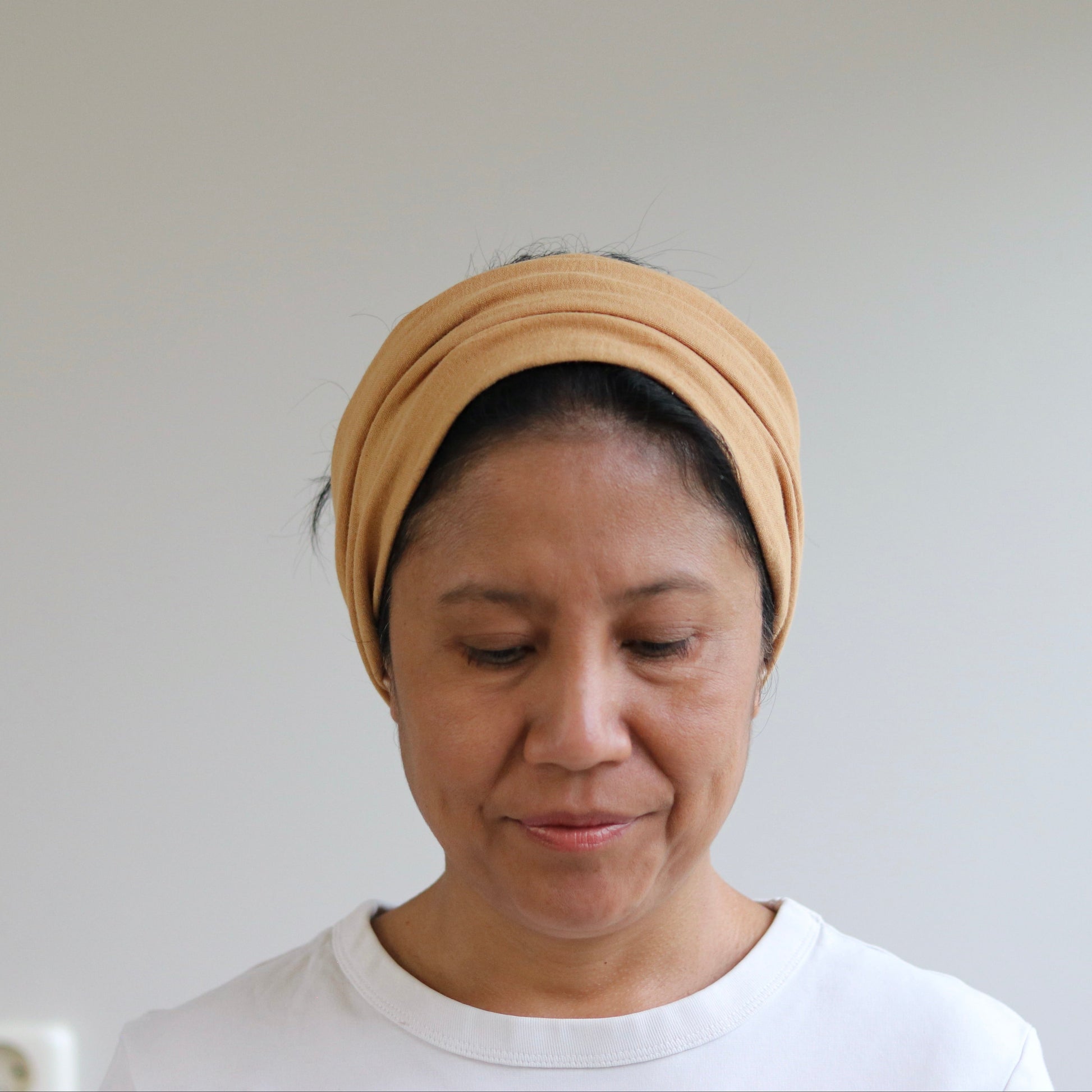 A woman wearing a caramel fabric headwrap or turban-style headband, paired with a white crewneck t-shirt, looking down against a plain light gray wall.