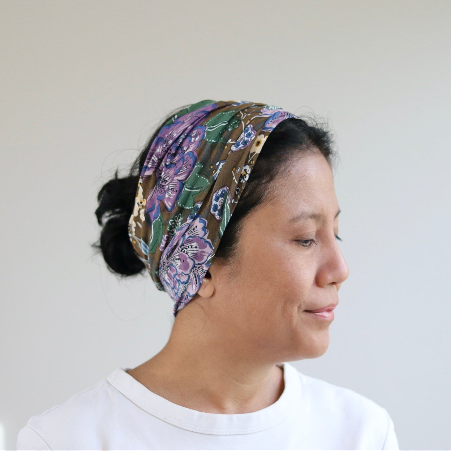 A woman in profile wearing a colorful patterned fabric headband and a white t-shirt against a plain light gray wall.