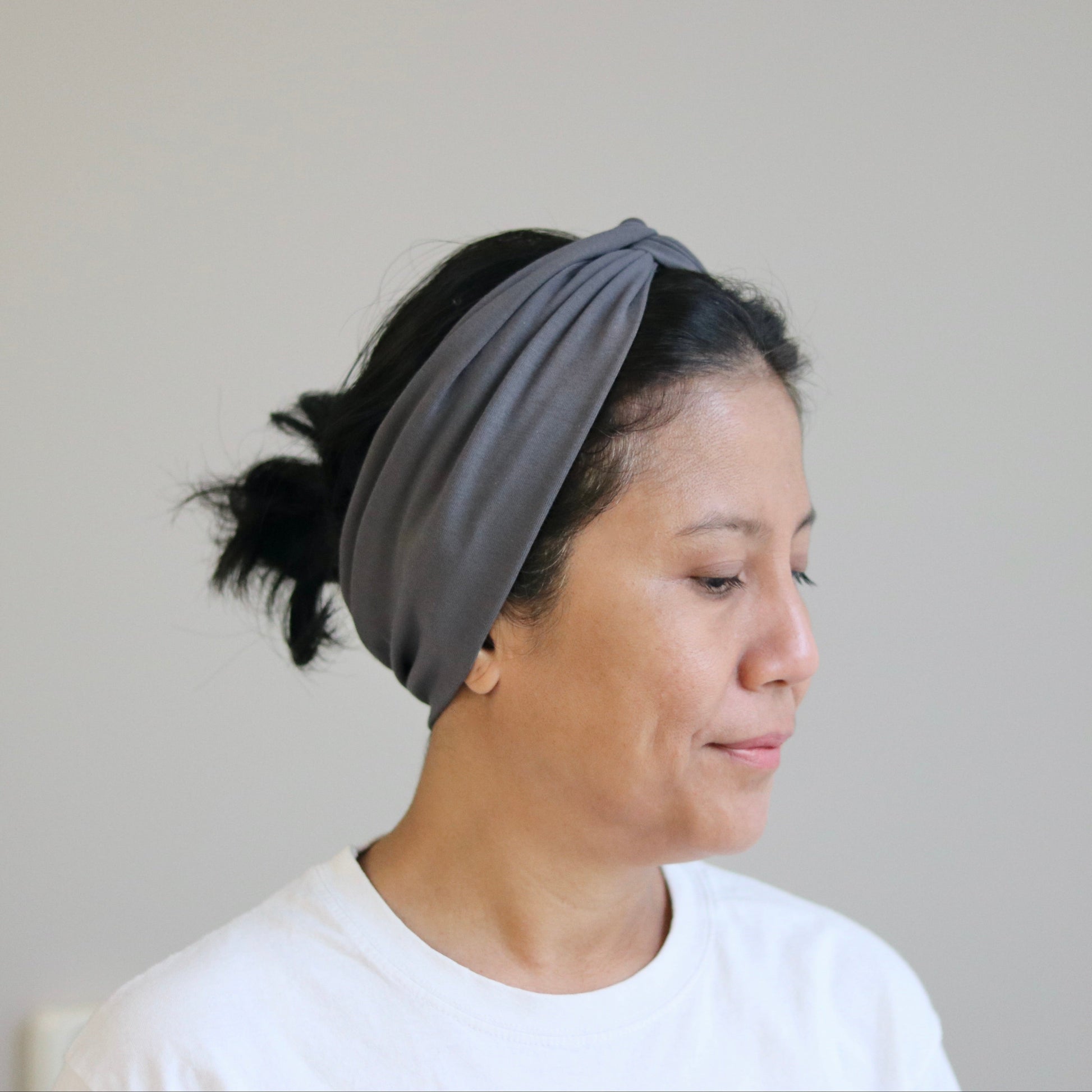 A woman wearing a dark gray knotted fabric headband and a white t-shirt, shown in profile against a plain light gray wall.
