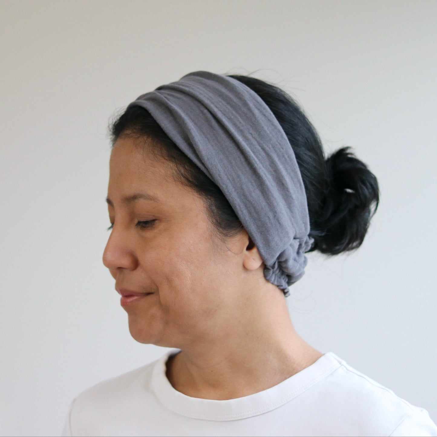 A woman wearing a wide, soft gray fabric headband with a knotted detail at the back of her dark hair, paired with a white t-shirt against a plain light gray wall.