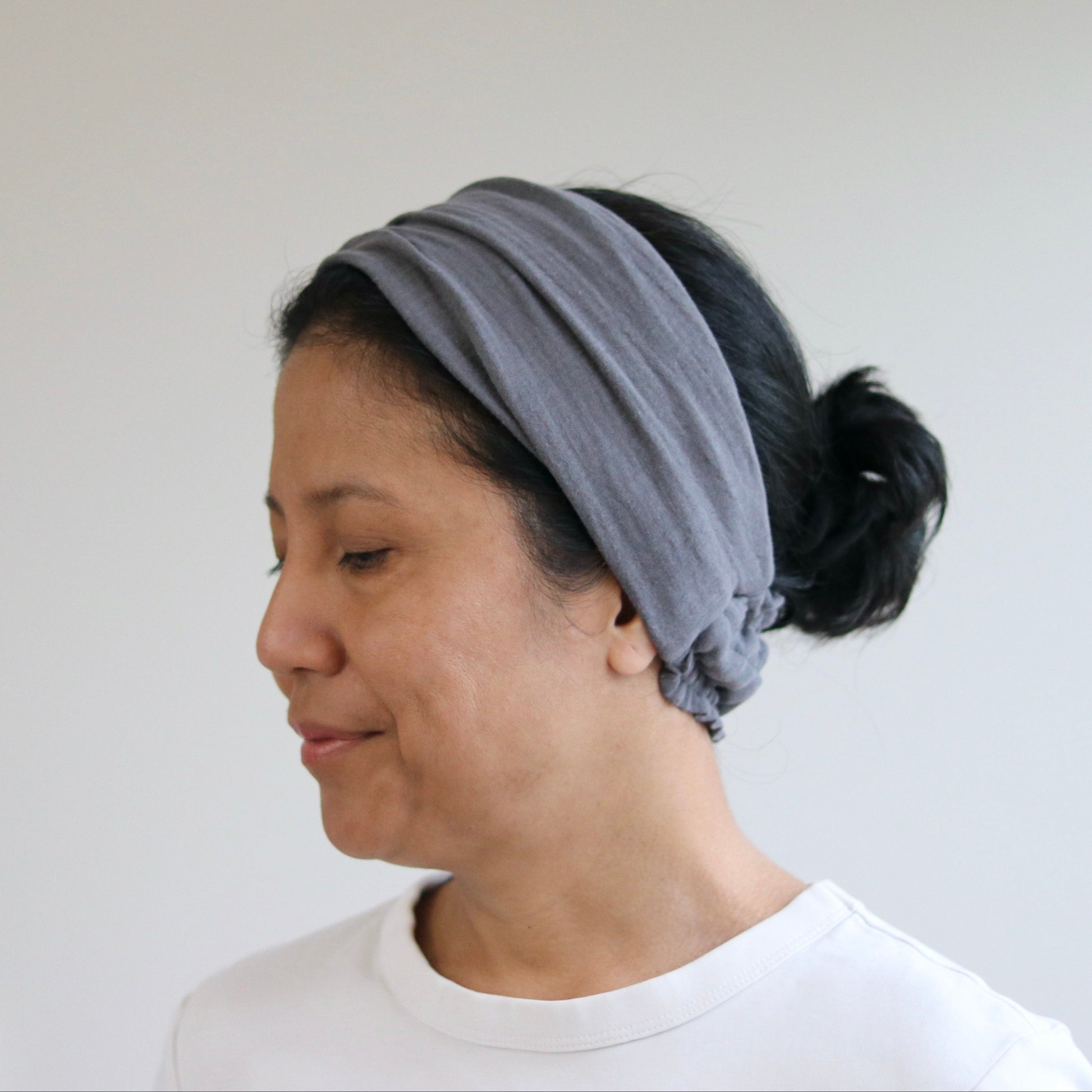 A woman wearing a wide, soft gray fabric headband with a knotted detail at the back of her dark hair, paired with a white t-shirt against a plain light gray wall.