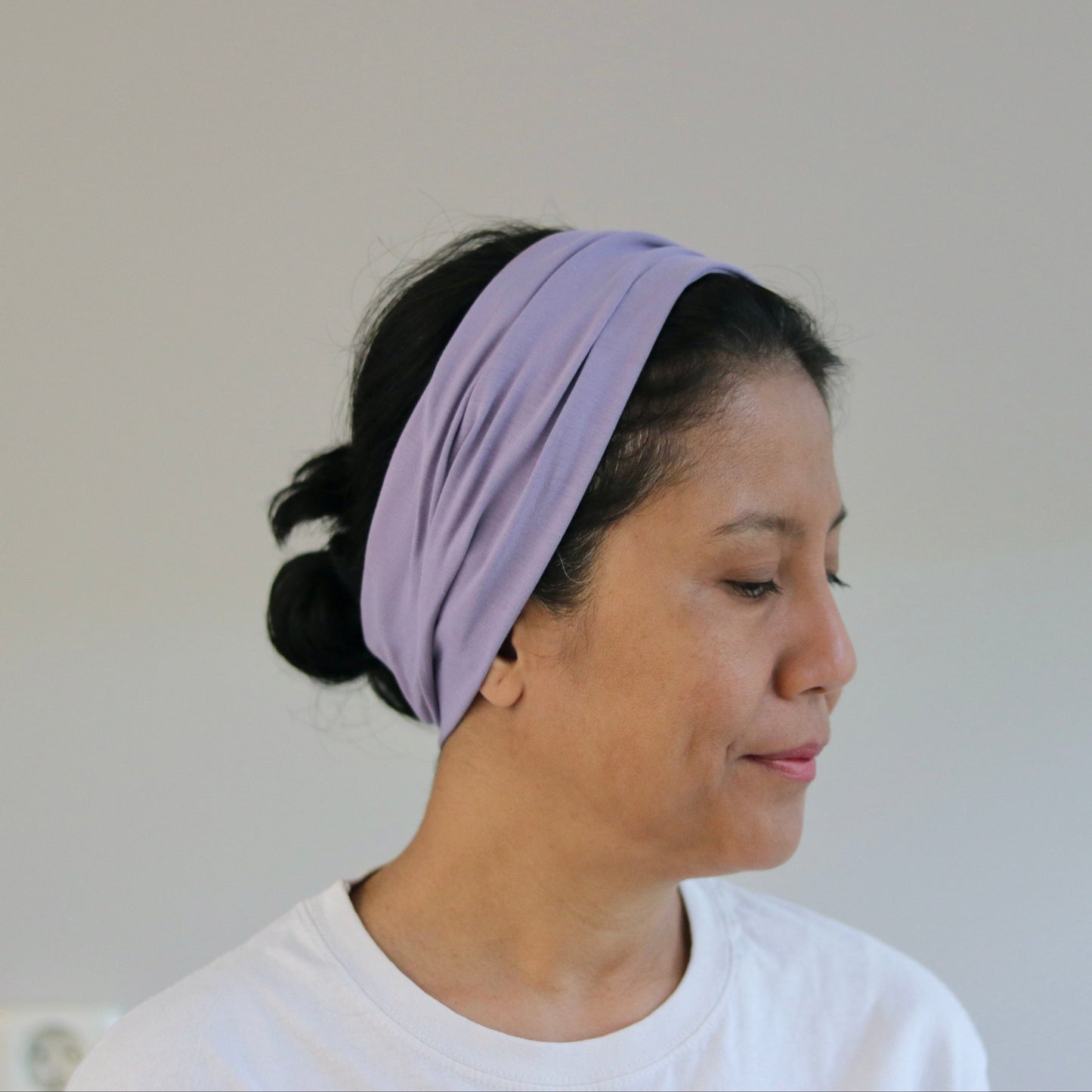 A woman wearing a light purple twisted fabric headband, shown in profile against a plain light gray wall.