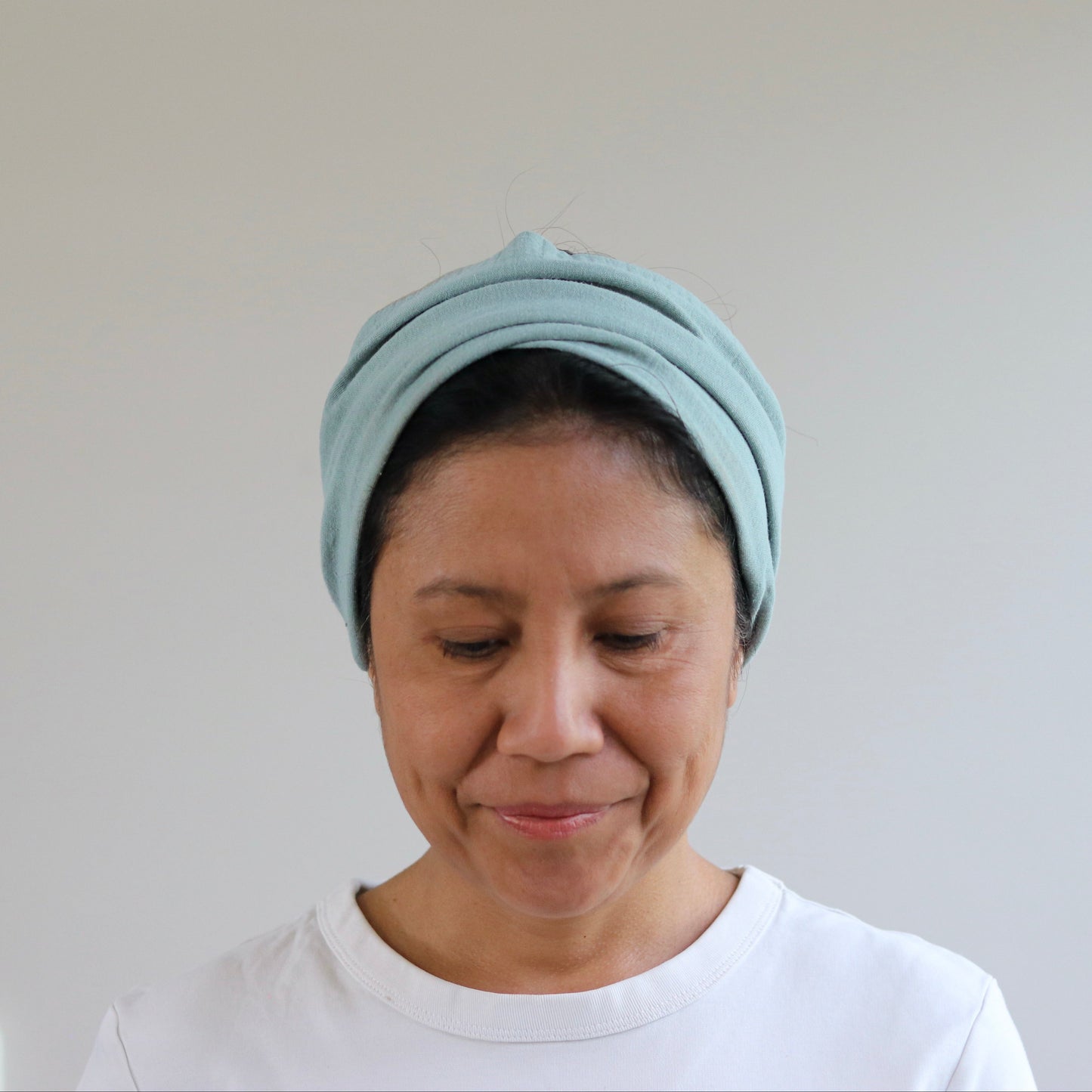 A woman wearing a dusty- mint knitted head wrap or turban-style headband, paired with a white t-shirt, looking down against a plain light gray wall background.