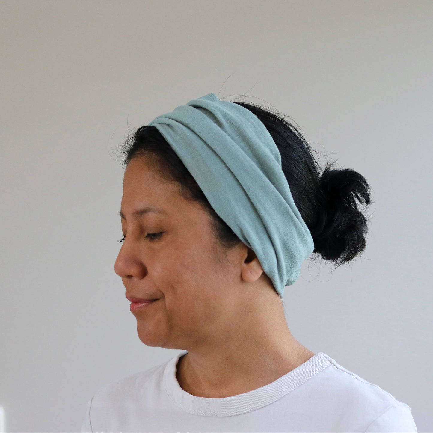 A woman wearing a dusty- mint fabric headband with her hair tied back in a bun, shown in profile against a plain light gray wall.