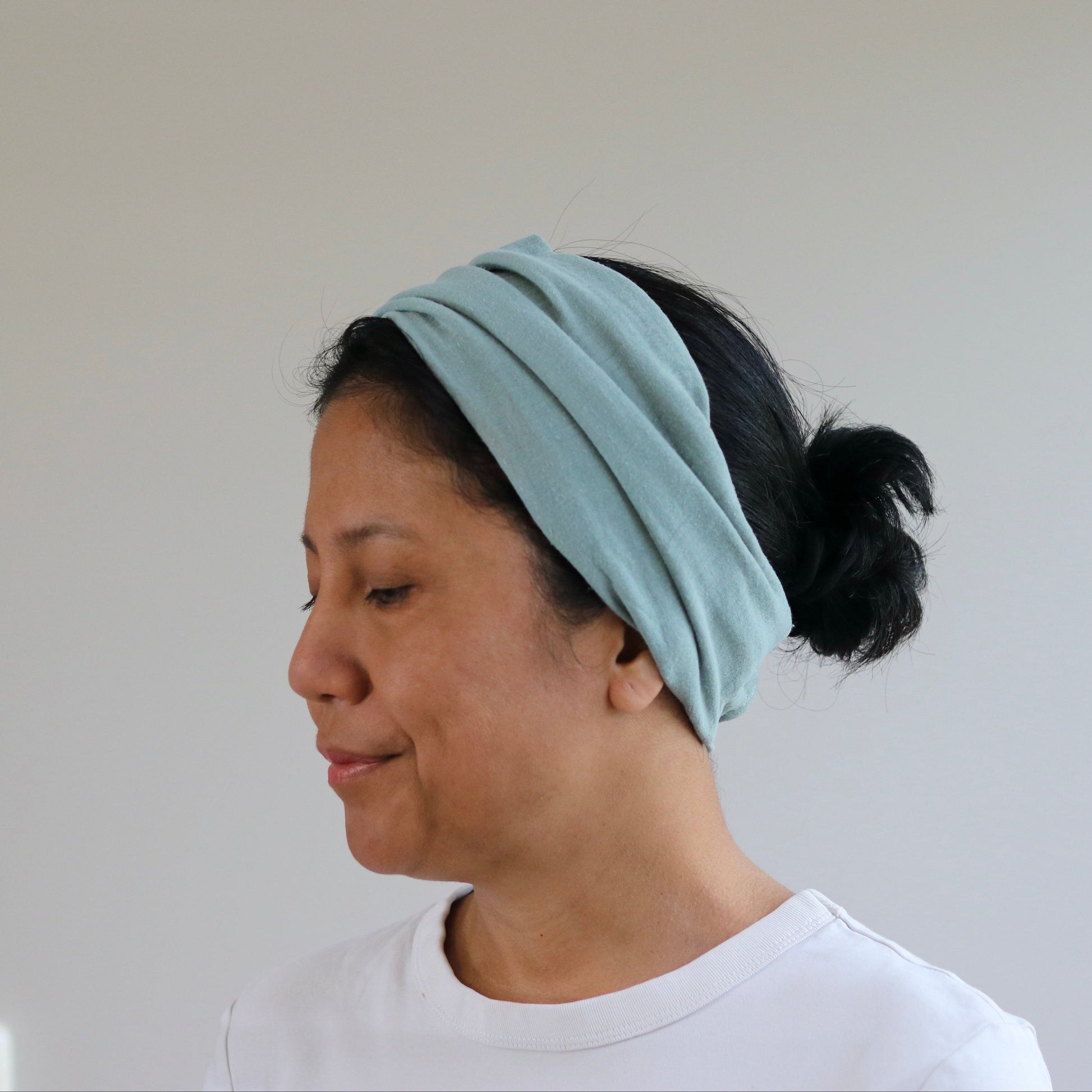 A woman wearing a dusty- mint fabric headband with her hair tied back in a bun, shown in profile against a plain light gray wall.
