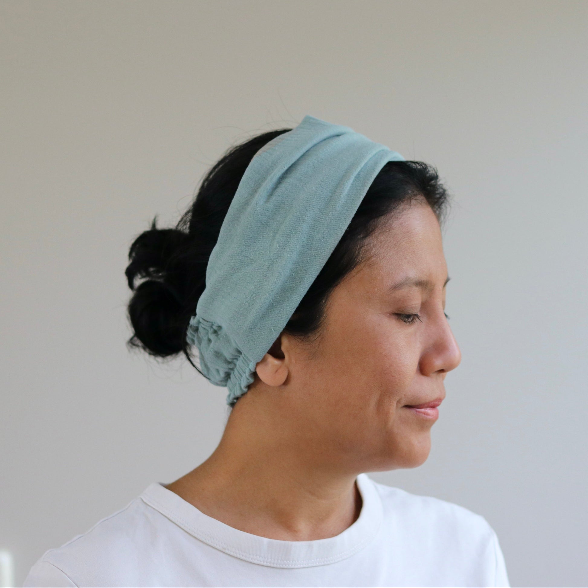 A woman in profile wearing a dusty- mint fabric headband and a white t-shirt, set against a plain light gray wall.