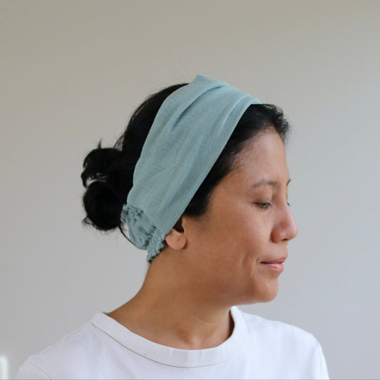 A woman in profile wearing a dusty- mint fabric headband and a white t-shirt, set against a plain light gray wall.