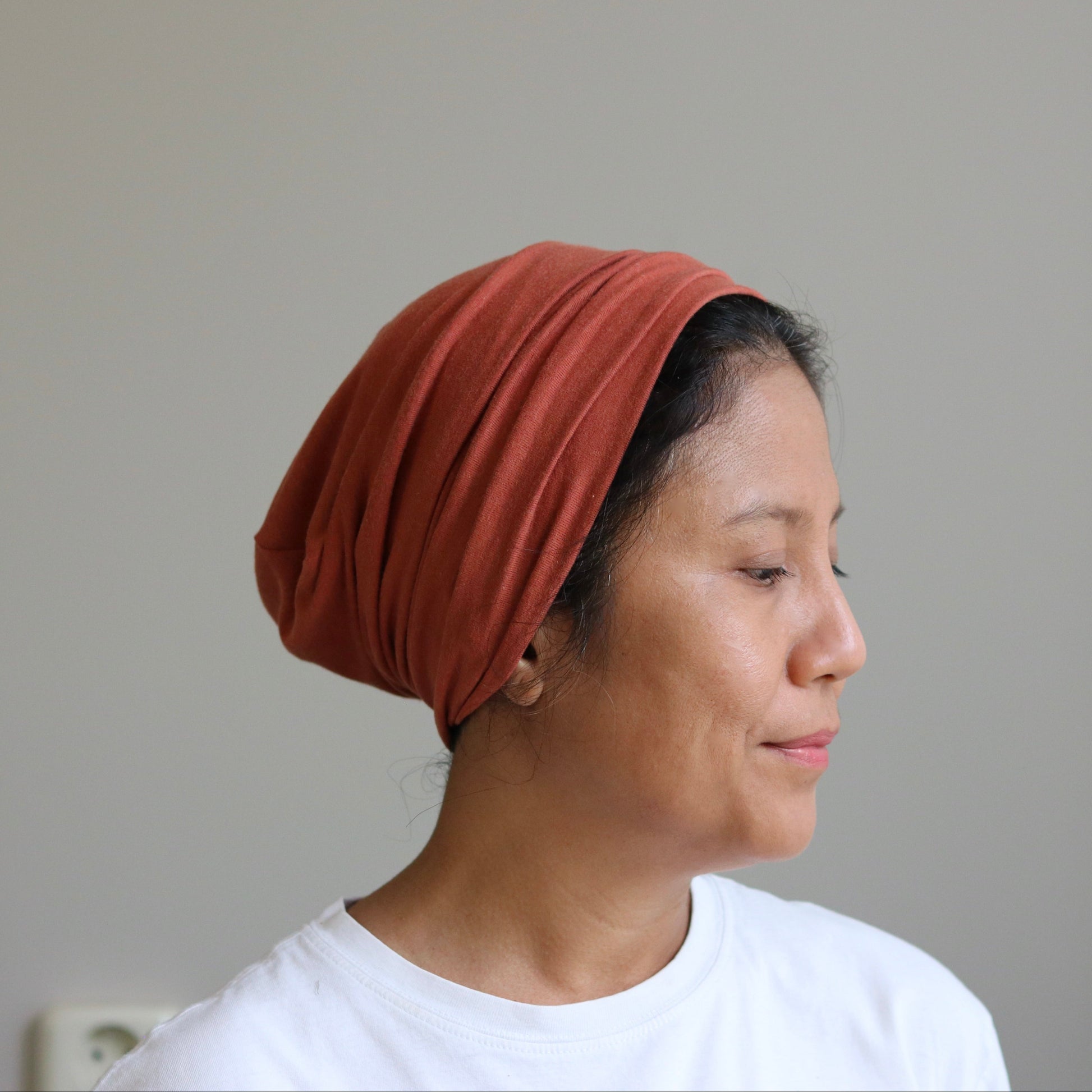 A woman in profile wearing a rust-colored, gathered fabric head covering or turban-style cap, paired with a white t-shirt, set against a plain light gray wall.