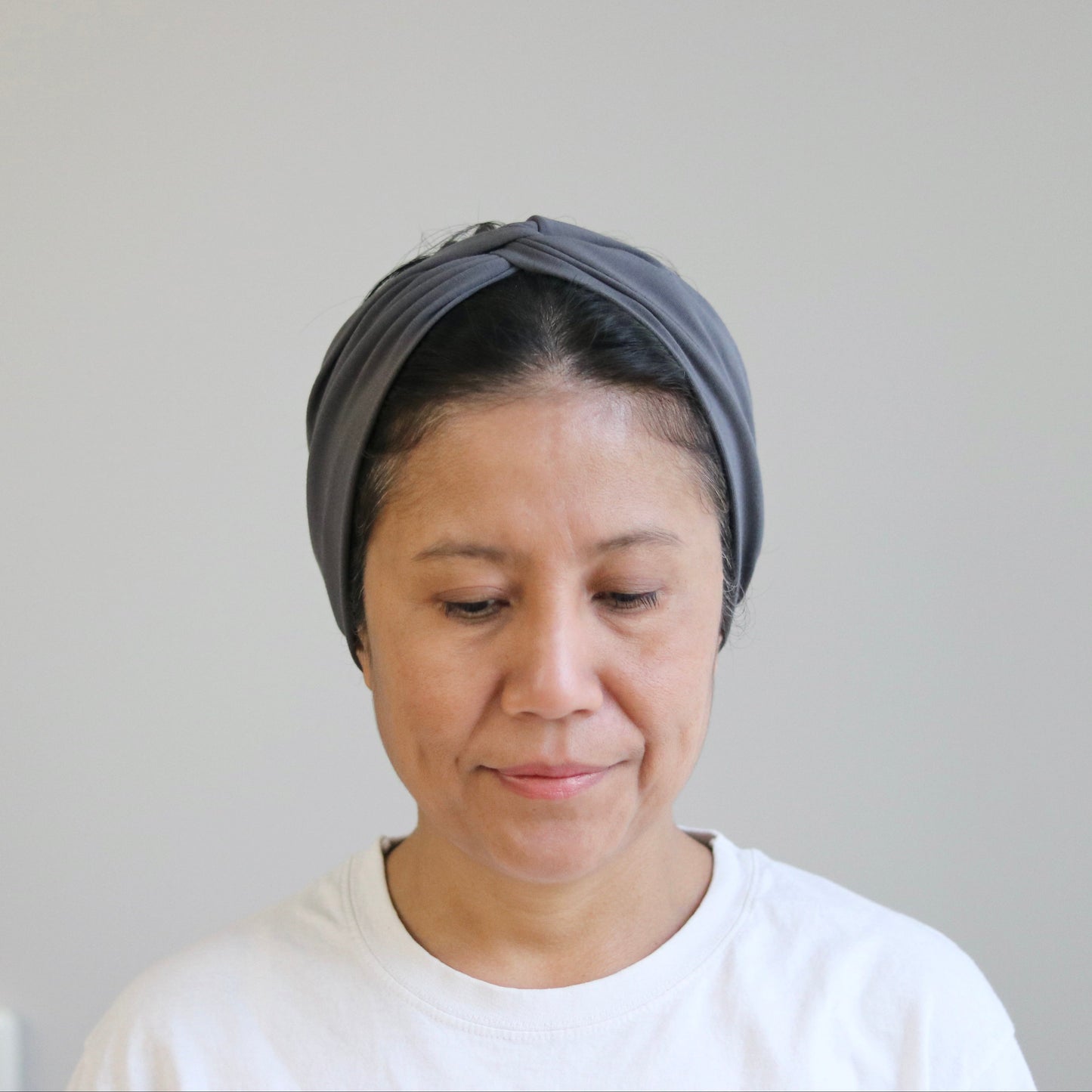 A woman wearing a dark gray twisted headband and a white t-shirt, looking down with a neutral expression against a plain light gray wall.