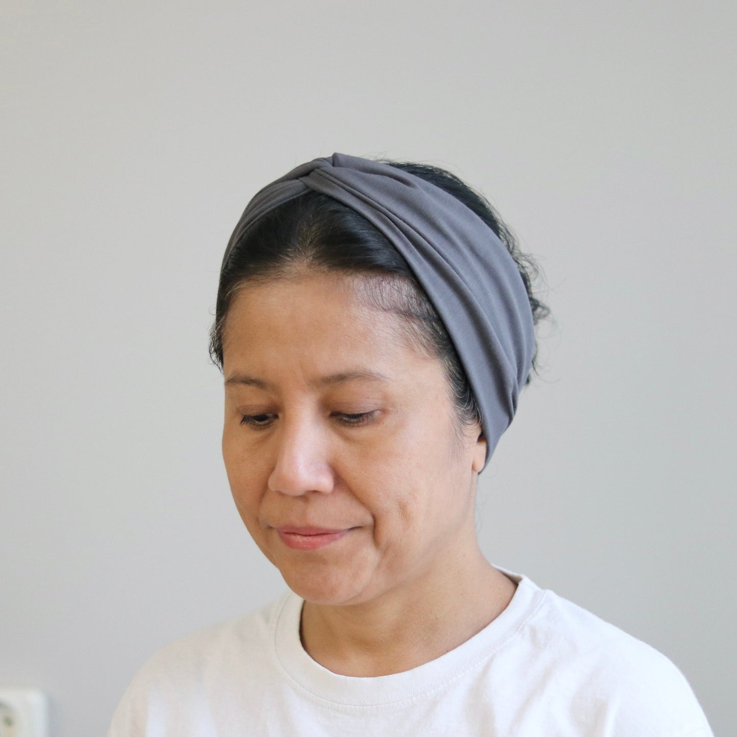 A woman wearing a gray fabric headband and a white t-shirt, looking down against a plain light gray wall background.
