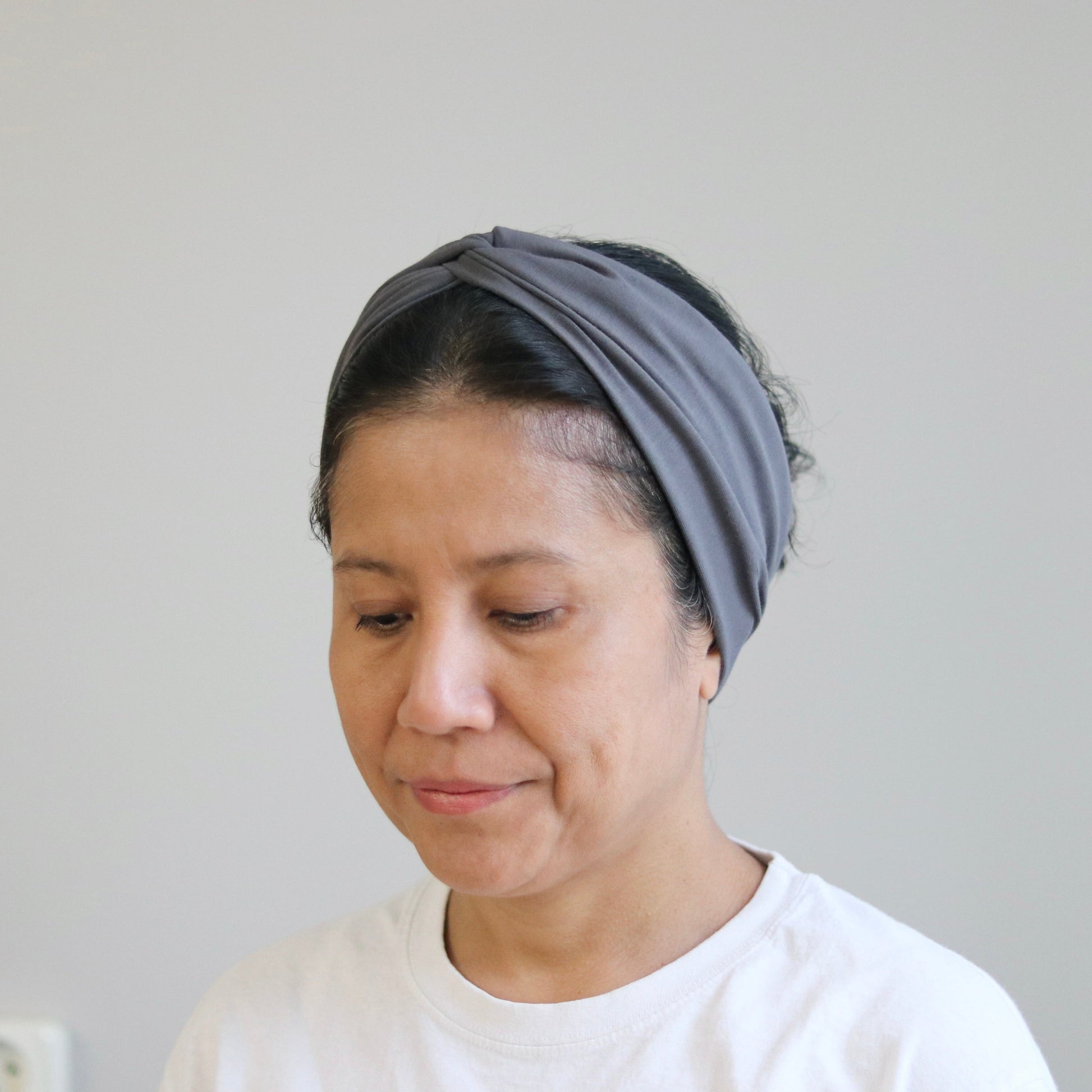 A woman wearing a gray fabric headband and a white t-shirt, looking down against a plain light gray wall background.