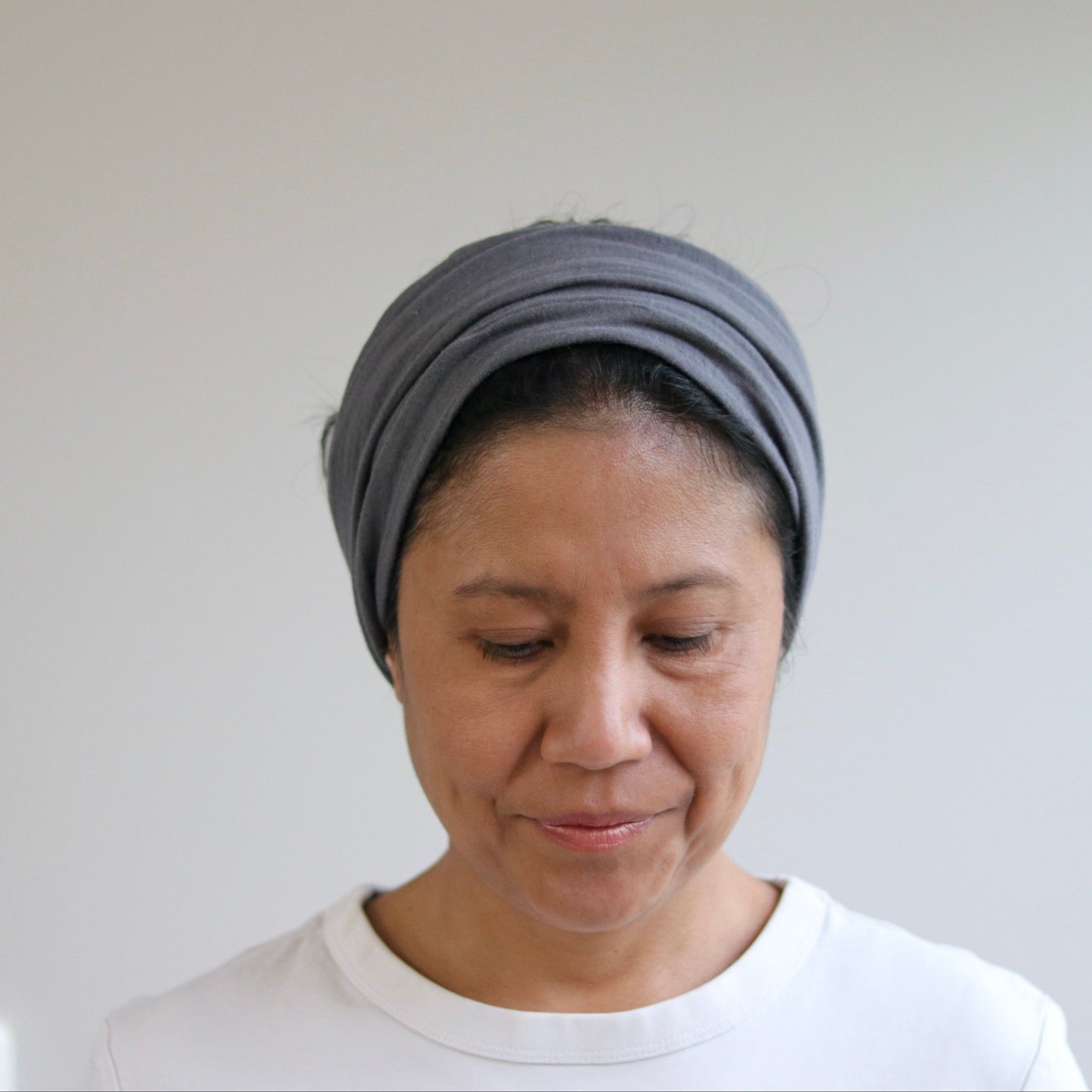 A woman wearing a gray fabric headwrap or turban-style hair accessory, paired with a white top, shown against a plain light gray wall.