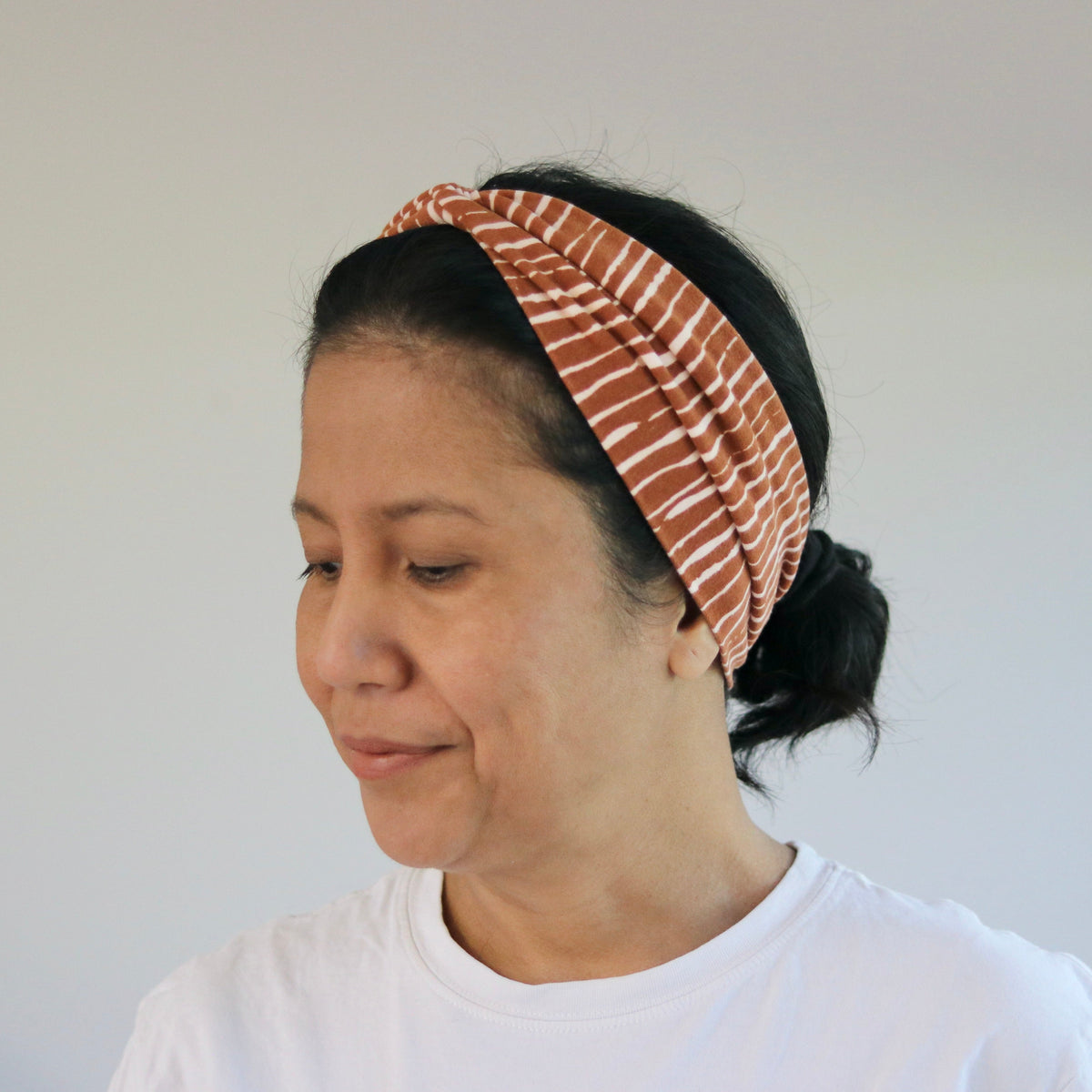 A woman wearing a rust-orange and white striped fabric headband, styled in a side profile view against a plain light gray wall.