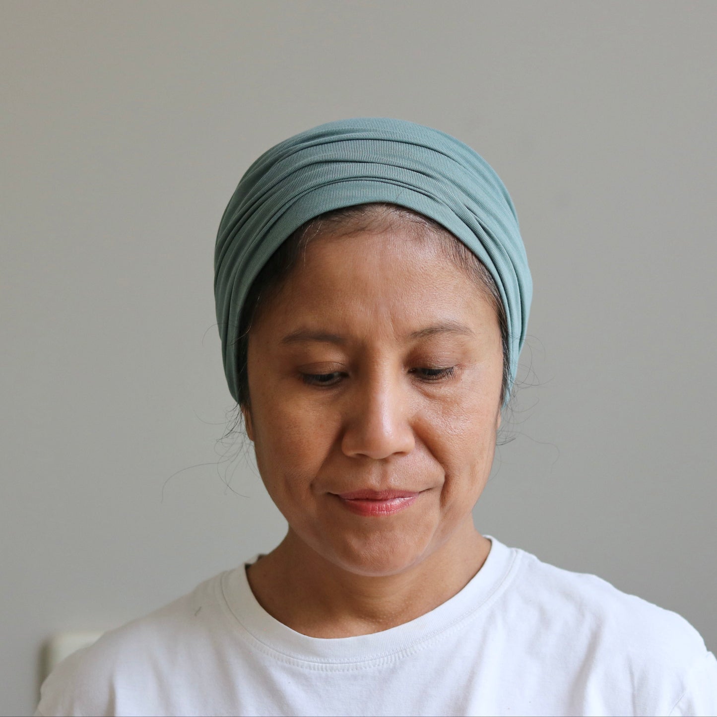 A woman wearing a light sage- green fabric headwrap turban and a white t-shirt, looking down against a plain light gray wall.