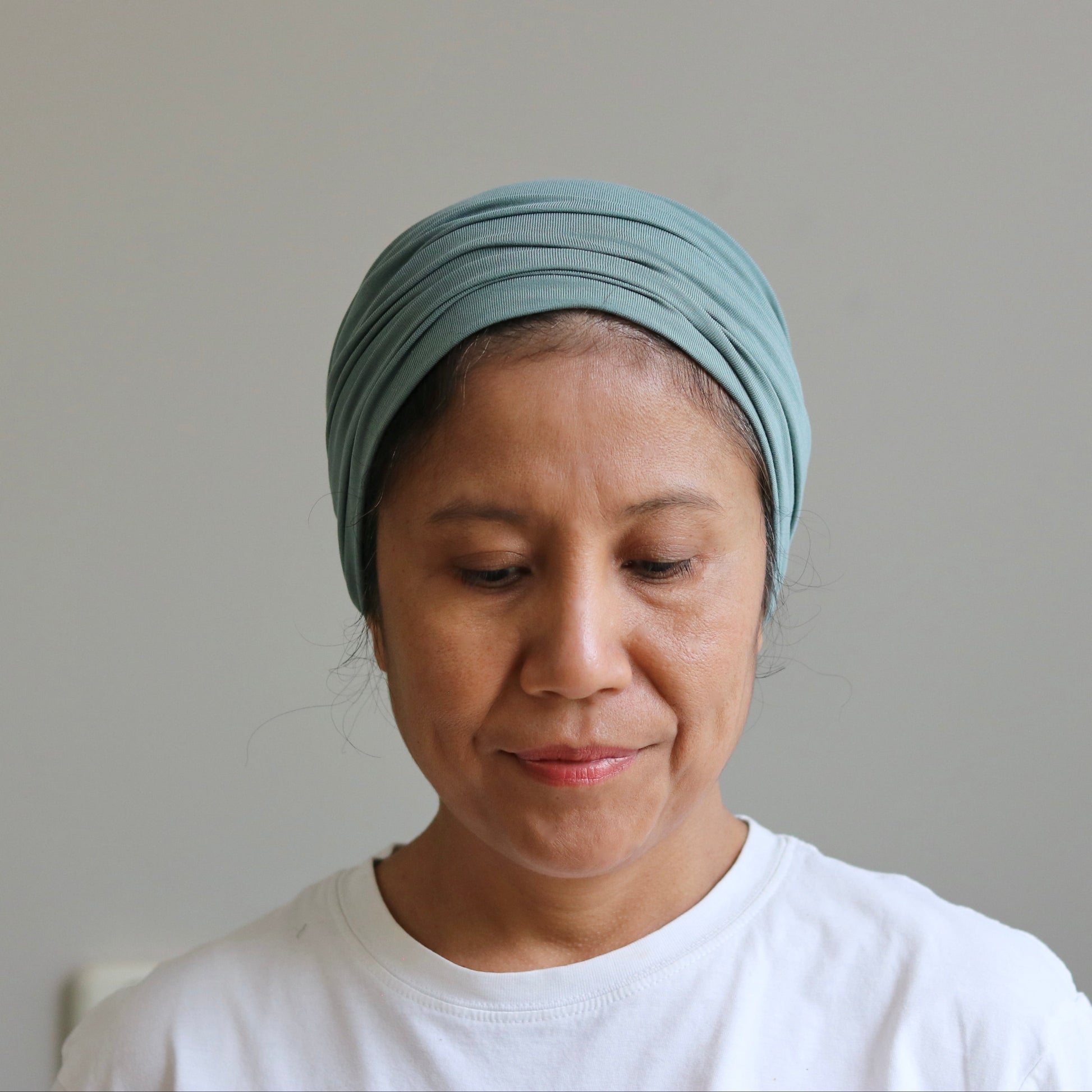 A woman wearing a light sage- green fabric headwrap turban and a white t-shirt, looking down against a plain light gray wall.