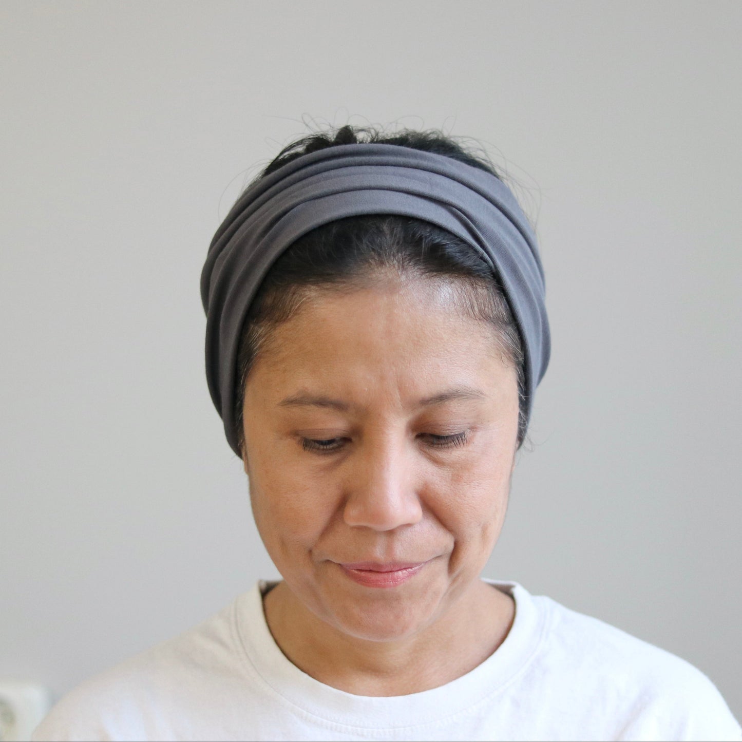 A woman wearing a gray multi-band headband and a white t-shirt, looking down against a plain light gray wall.
