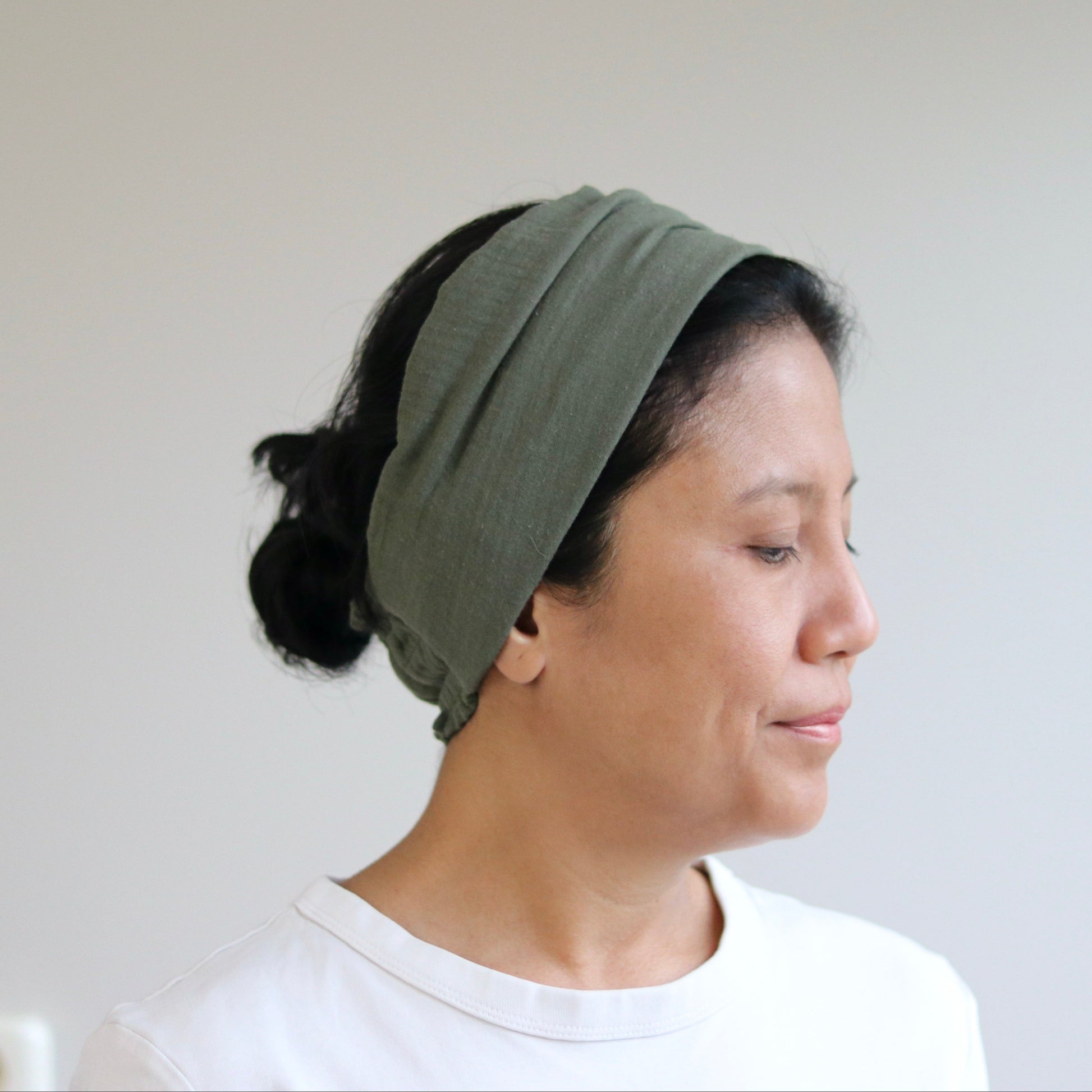 A woman in profile wearing an olive green fabric headband and a white t-shirt, set against a plain light gray wall.