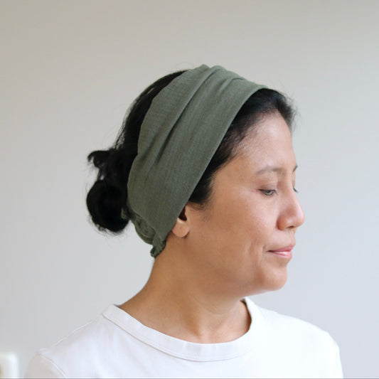 A woman in profile wearing an olive green fabric headband and a white t-shirt, set against a plain light gray wall.
