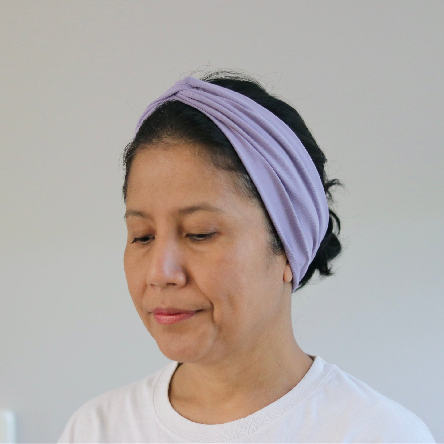 A woman wearing a light purple twisted fabric headband and a white t-shirt, shown from the chest up against a plain light gray wall.