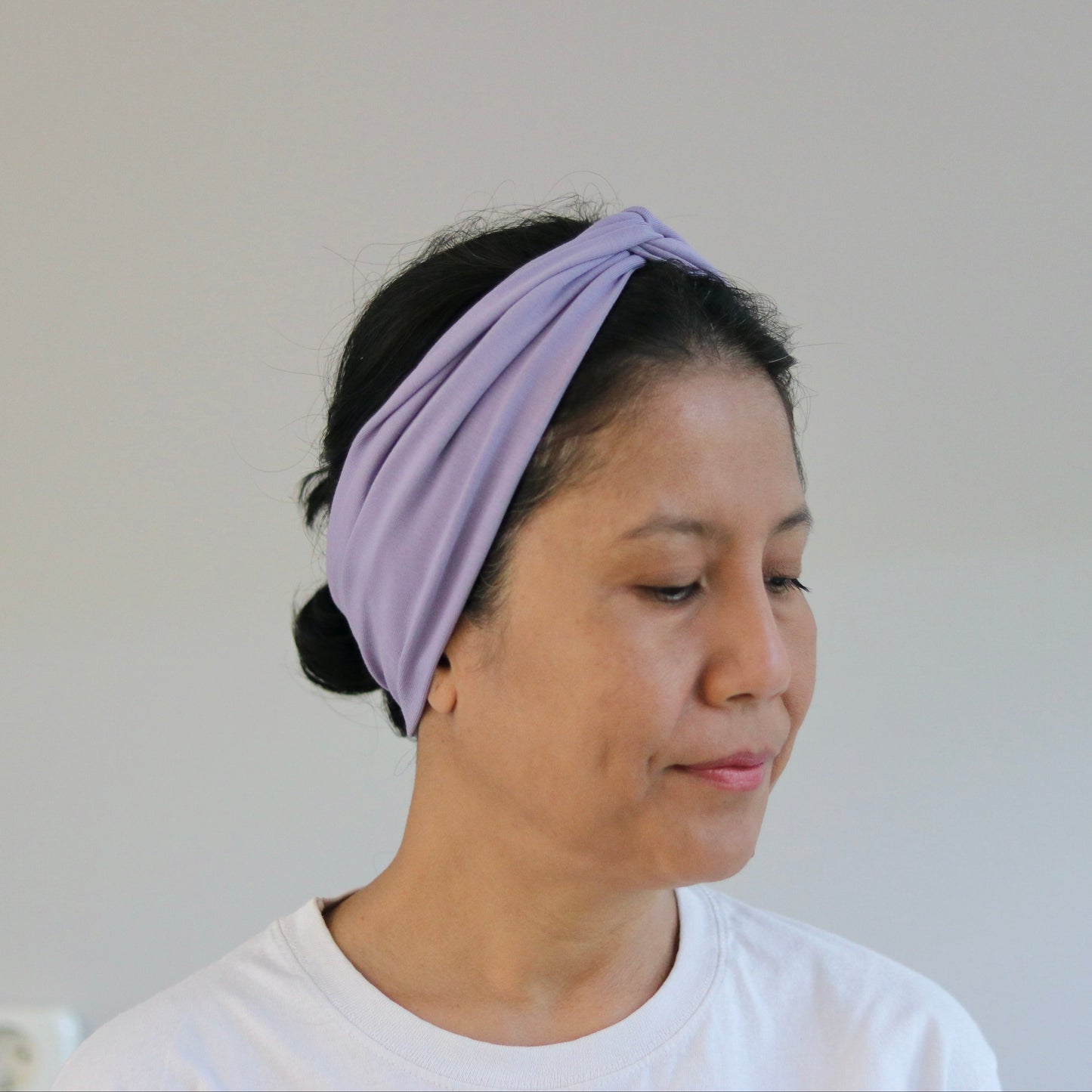 A woman wearing a light purple twisted fabric headband and a white t-shirt, shown from the chest up against a plain light gray wall.