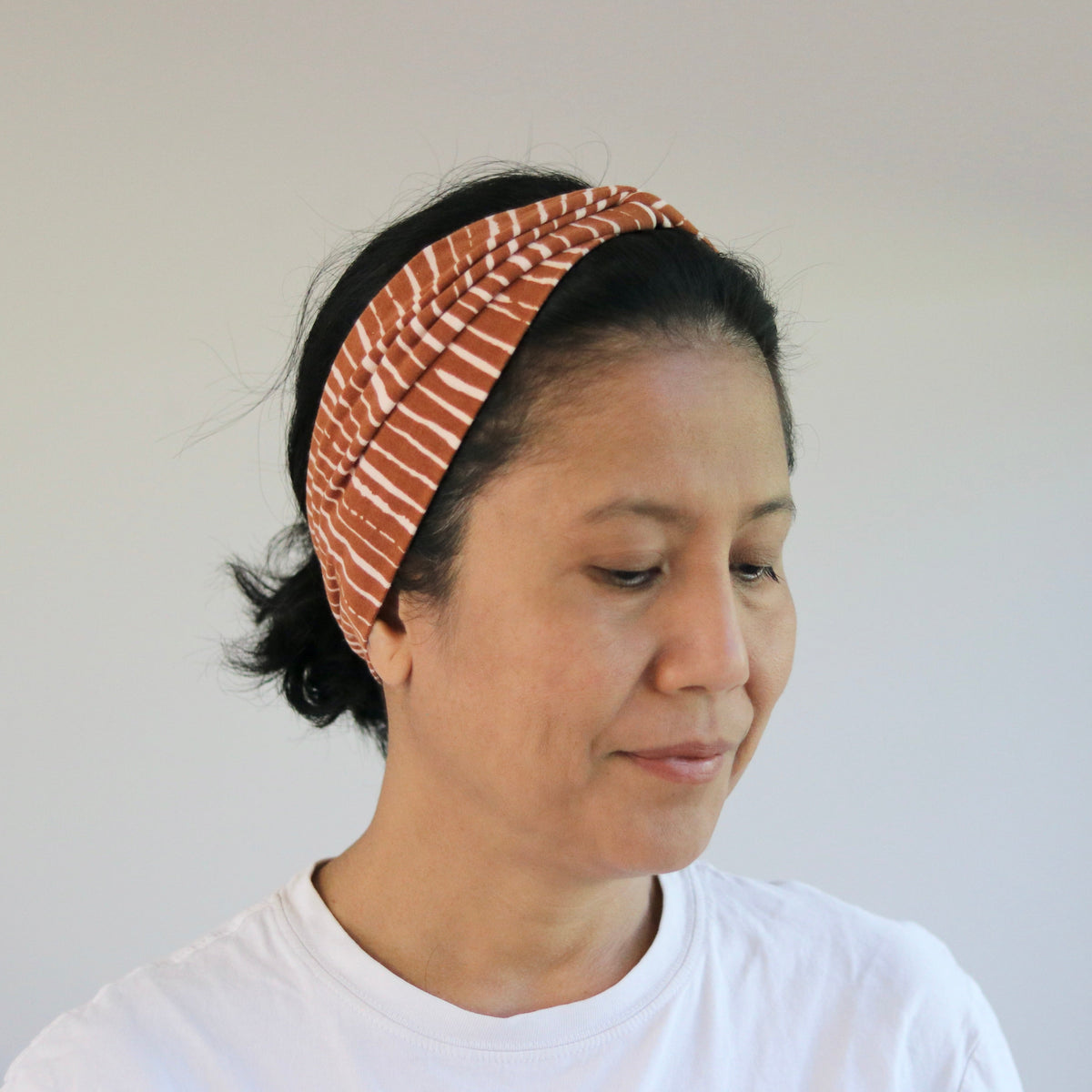 A woman wearing a rust-orange and white striped fabric headband, paired with a white t-shirt, photographed in a brightly lit indoor setting.