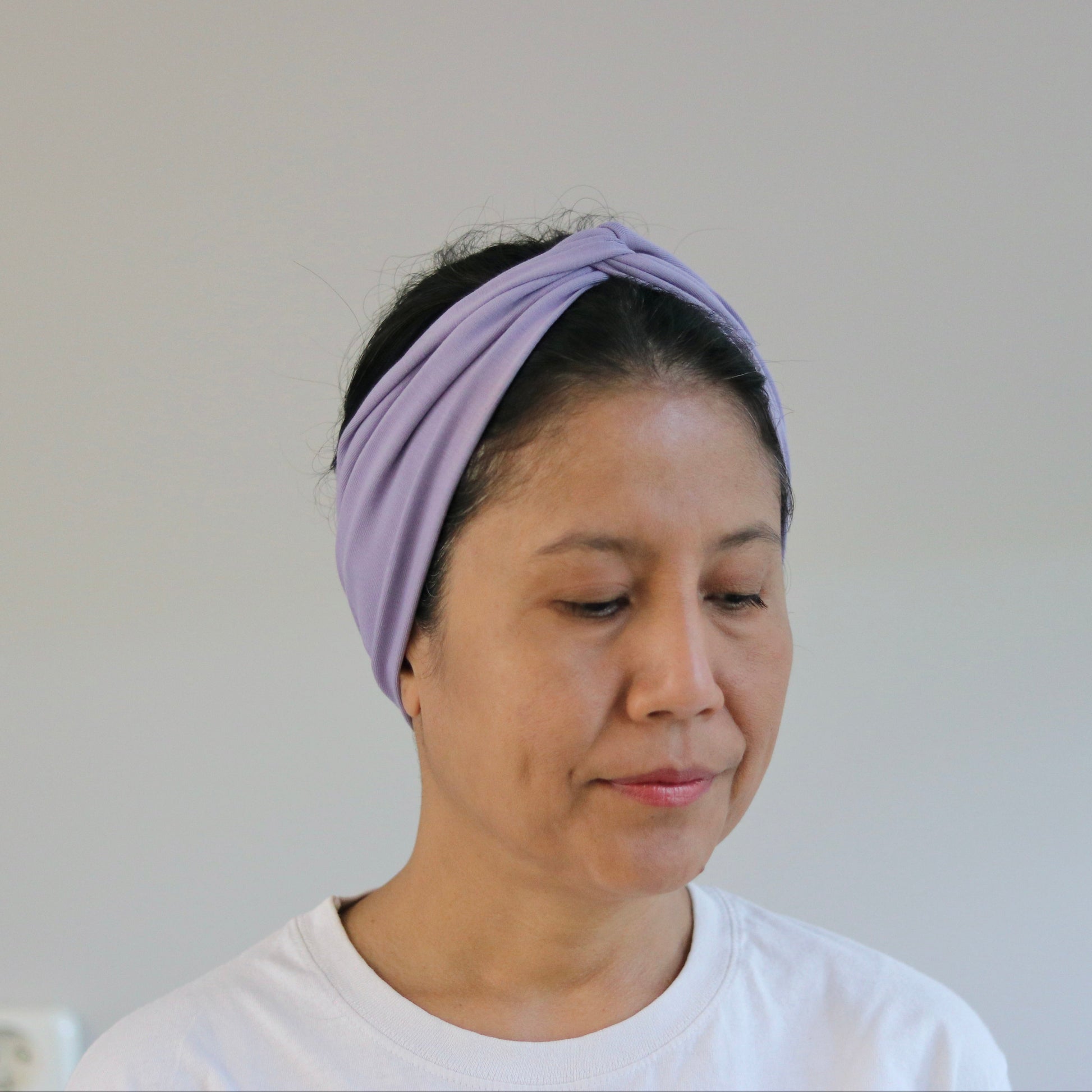 A woman wearing a light purple twisted fabric headband and a white t-shirt, shown from the chest up against a plain light gray wall.