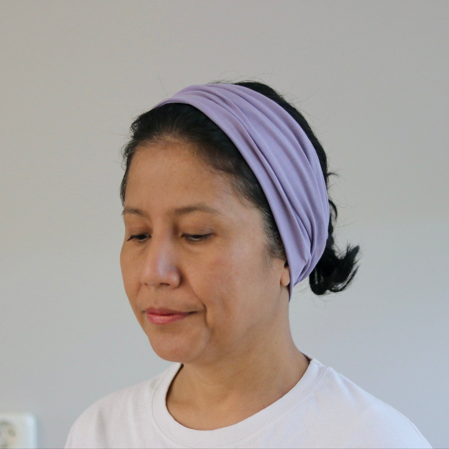 A woman wearing a light purple/lavender fabric headband and a white t-shirt, shown from the chest up against a plain light gray wall.
