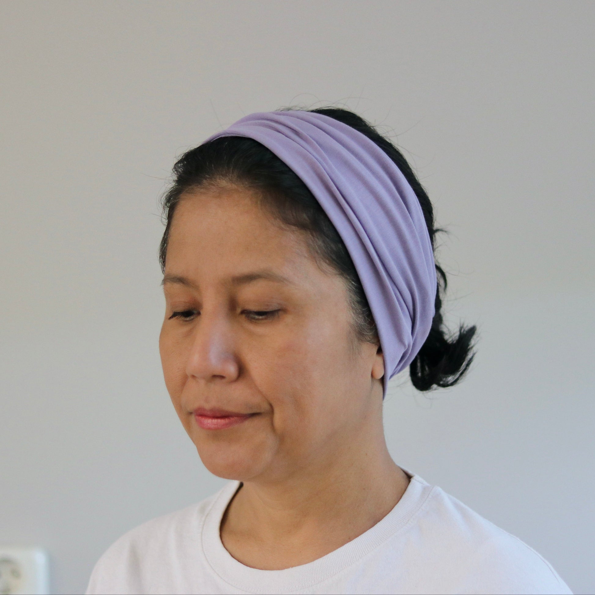 A woman wearing a light purple/lavender fabric headband and a white t-shirt, shown from the chest up against a plain light gray wall.