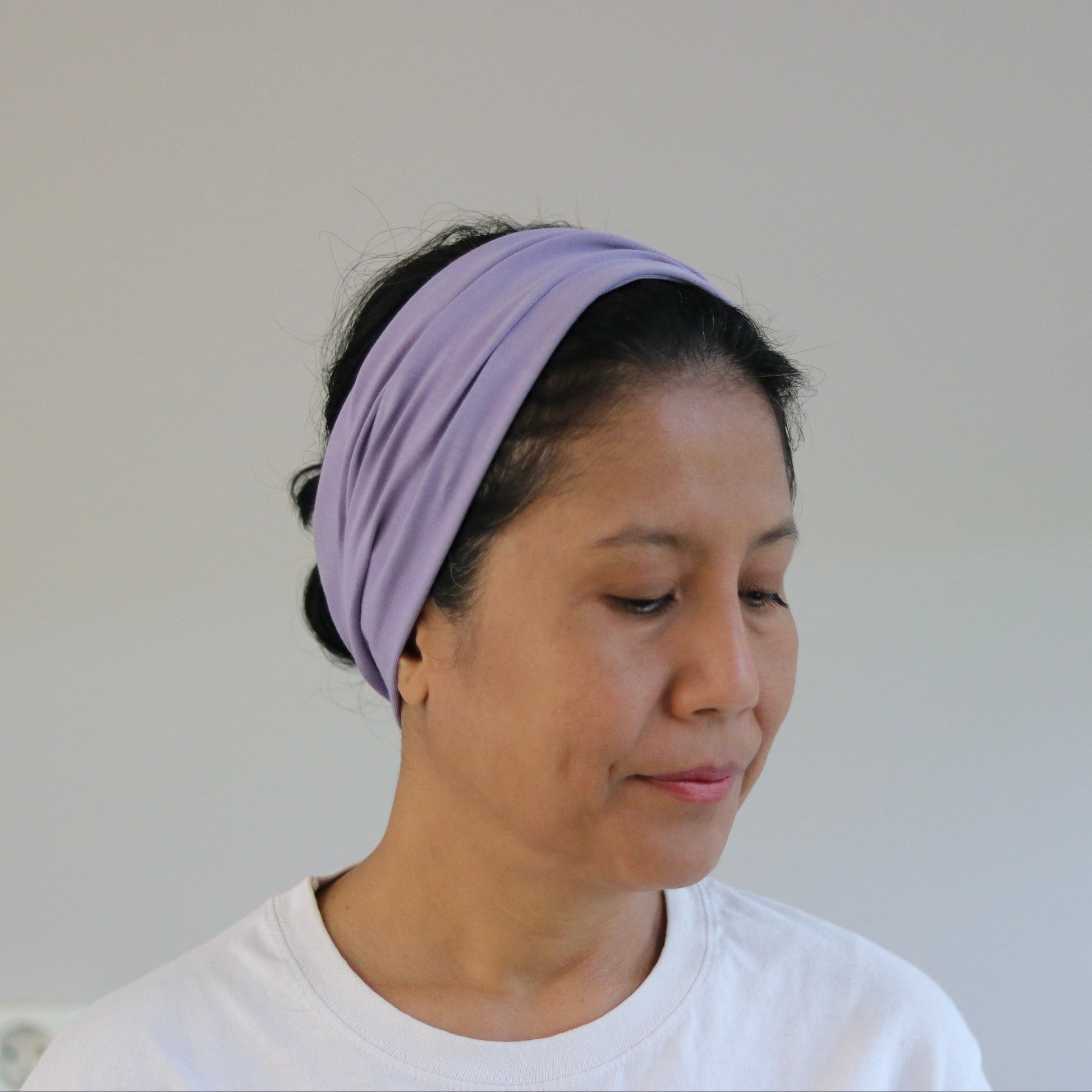 A woman wearing a light purple fabric headband and a white t-shirt, shown from the chest up against a plain light gray wall.
