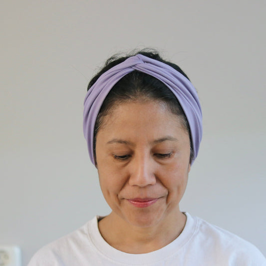 A woman wearing a light purple twisted fabric headband and a white t-shirt, looking down with a neutral expression against a plain light gray wall.