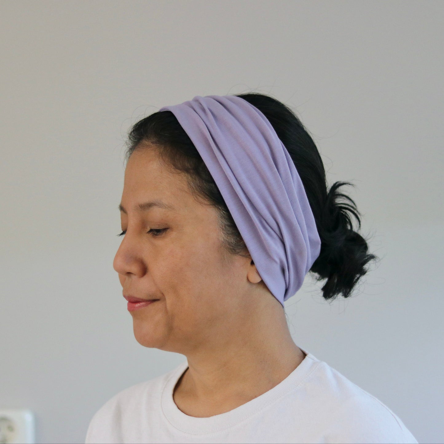 A woman wearing a light purple knotted fabric headband, shown in profile against a plain light gray wall.
